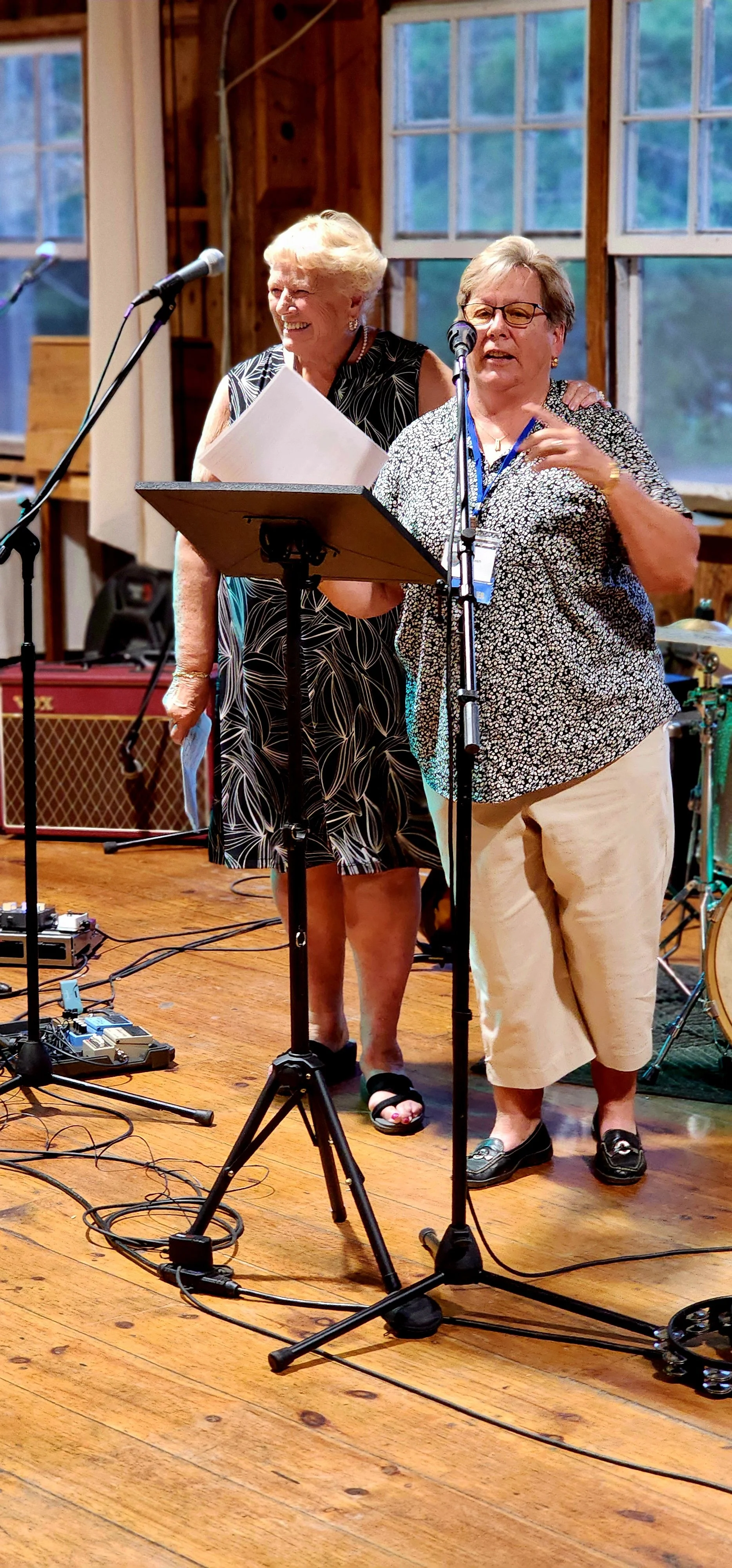 Two women, ones wearing a black dress with white patterns the other wearing a blue and white floral shirt with white pants, with a stand in front of them, one speaking into a microphone