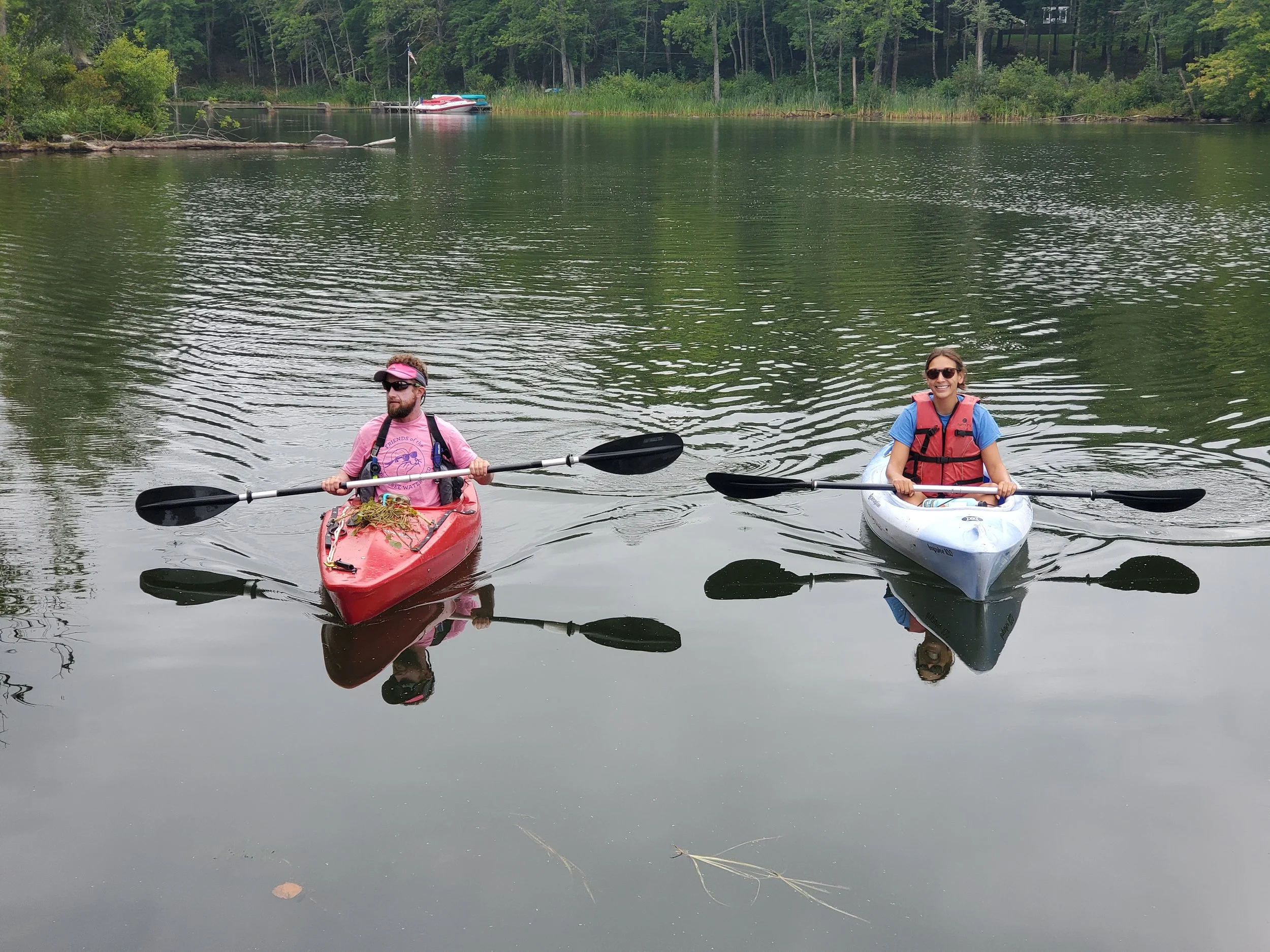 Two people in kayaks, the left in a red kayak wearing a pink t-shirt, black life vest, and sunglasses looking away from the camera, the one on the right in a blue kayak wearing a blue t-shirt, red life vest and smiling at the camera