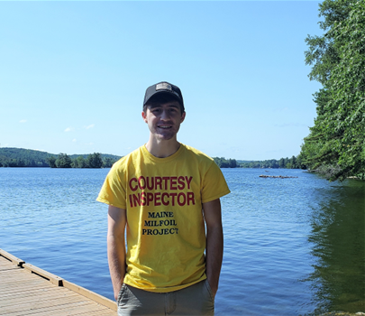 A man wearing a blue ball cap, yellow "Courtesy Inspector" shirt, smiling at the camera on a dock with a lake in the background