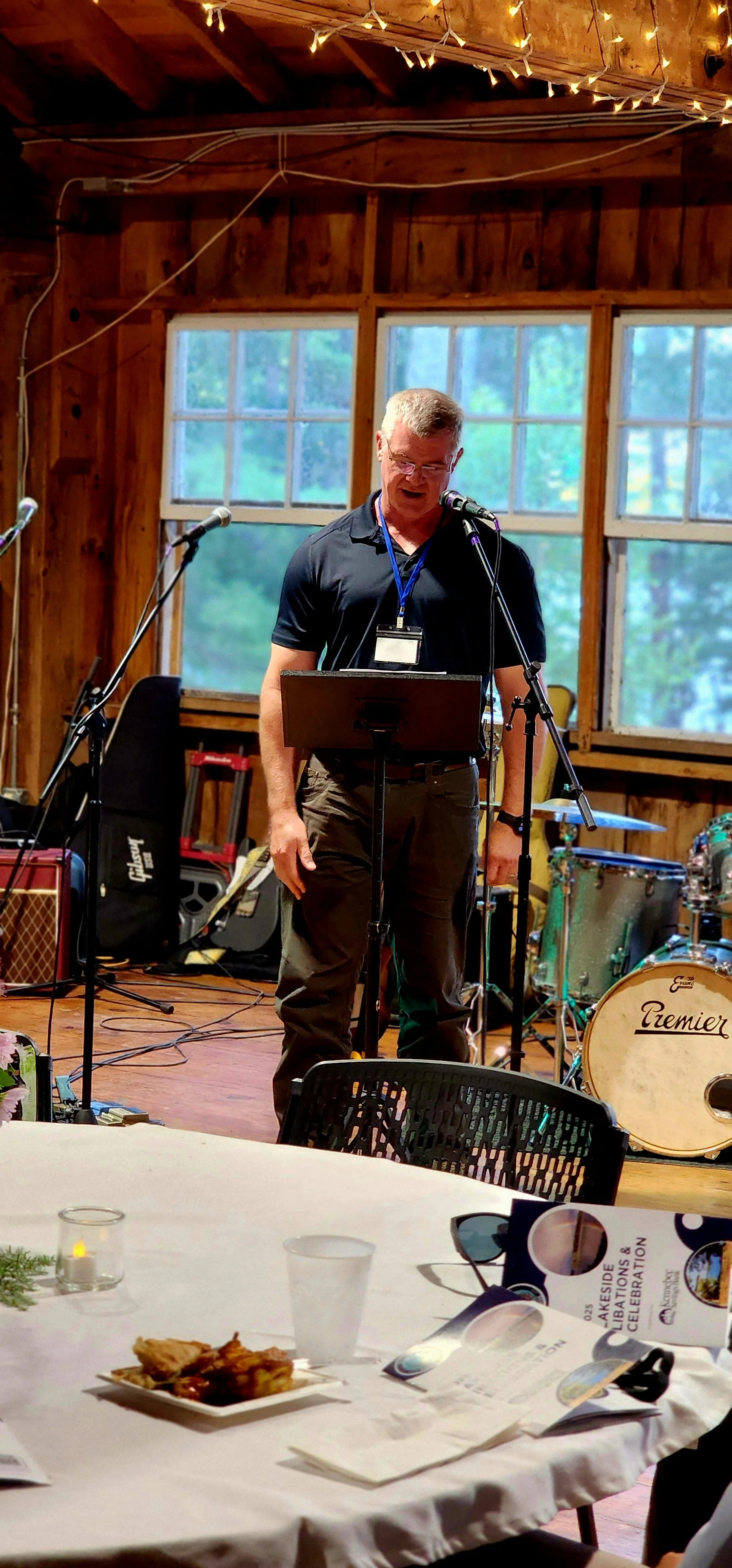 A man, wearing a black polo and dark grey pants, speaking into a microphone with a stand in front of him