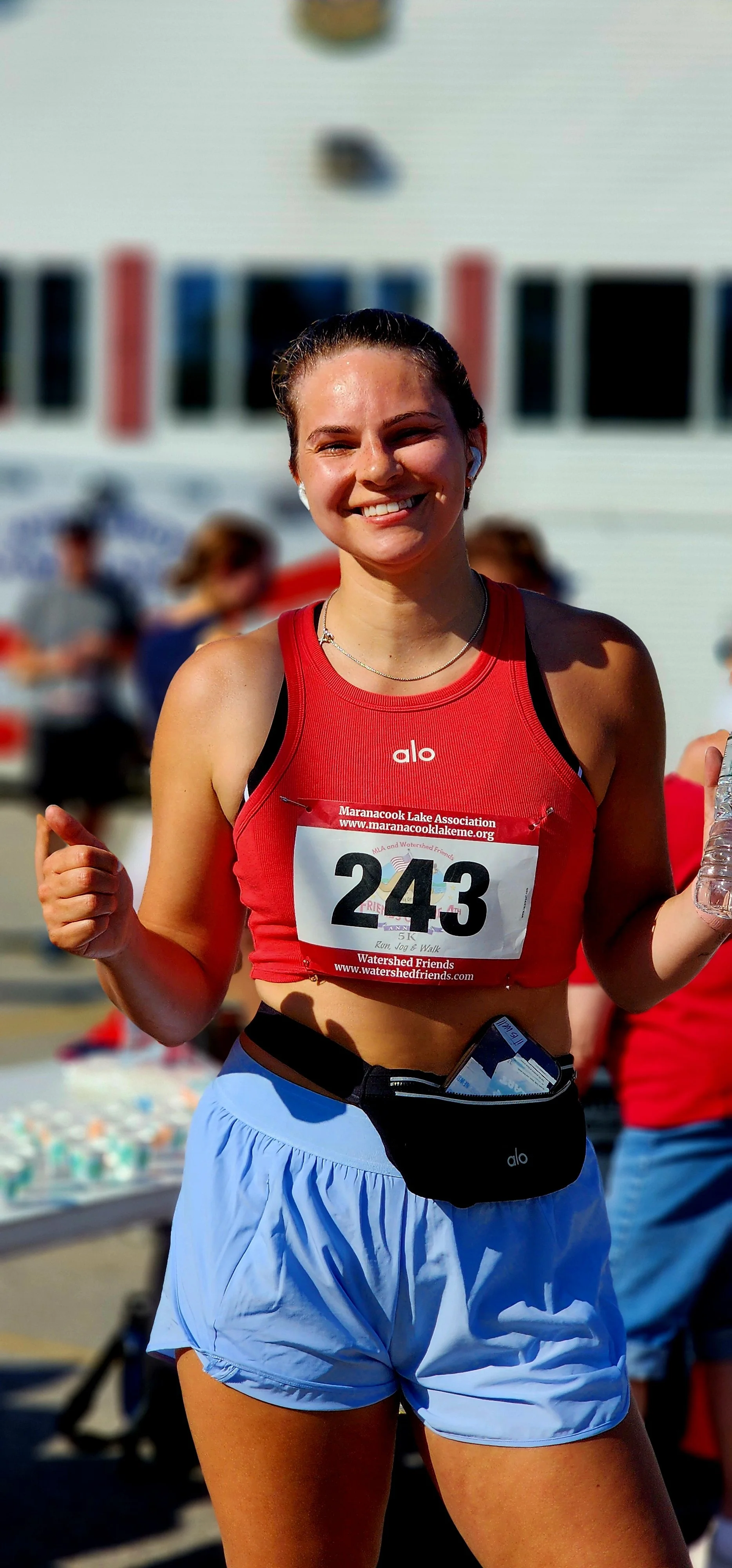 A woman wearing a red cropped tank top and blue shorts smiling and giving the camera a thumbs up
