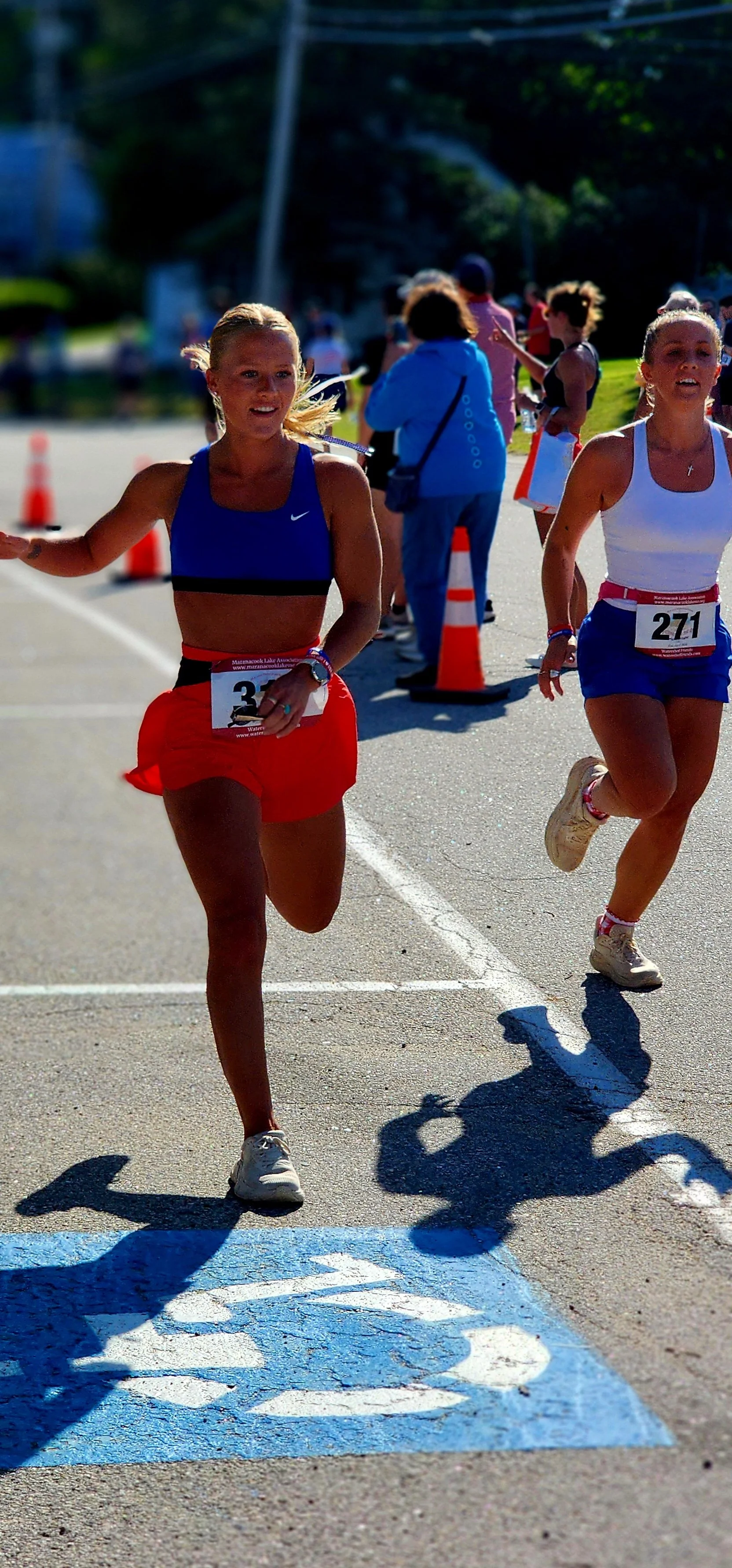 Two women running, one wearing a blue sports bra and red shorts the other wearing a white tank top and blue shorts