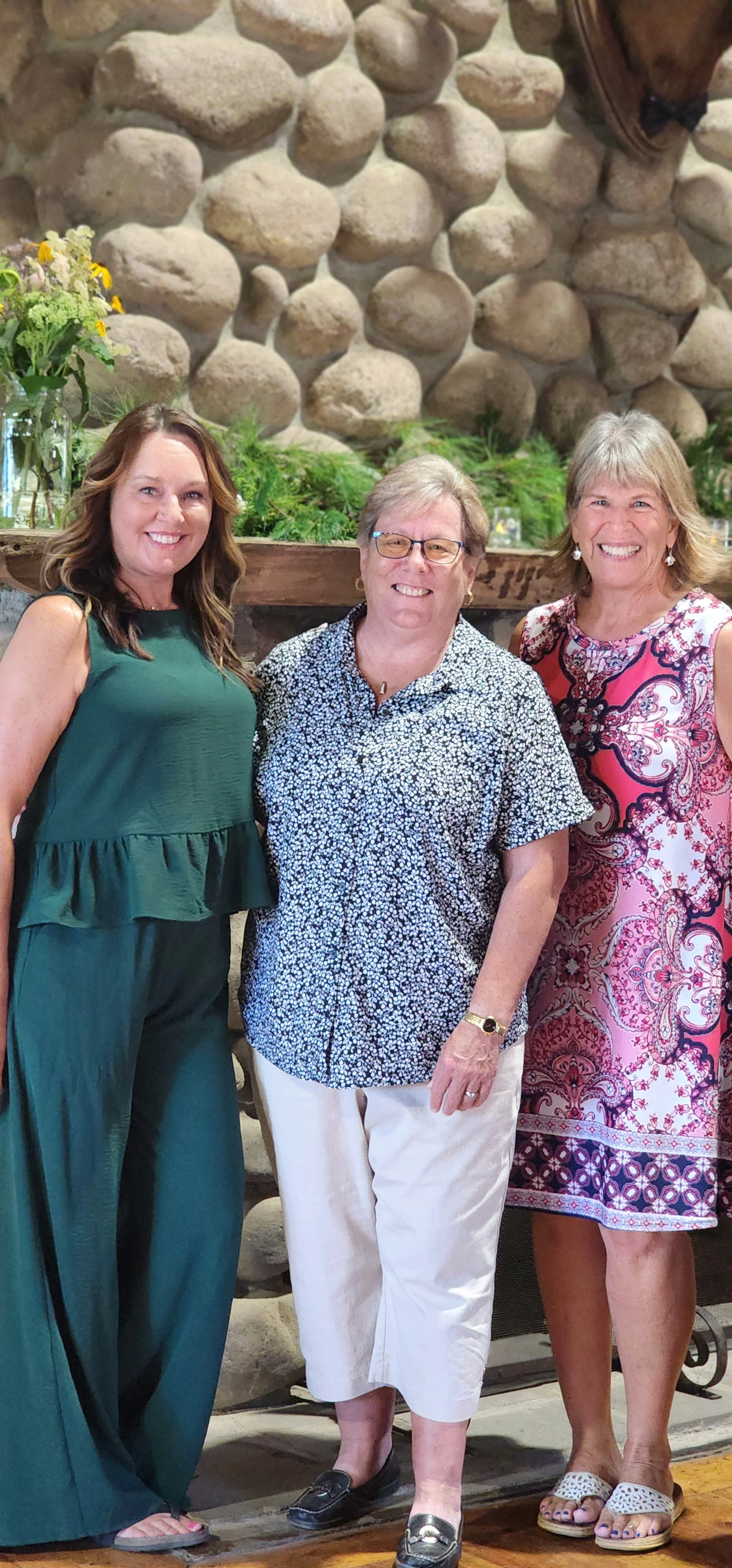 Three women, the one on the left wearing a green jump suit, the one in the middle wearing a white and blue patterned shirt and white pants and the one on the right wearing a pink paisley pattern dress, standing in front of a fireplace that has greene
