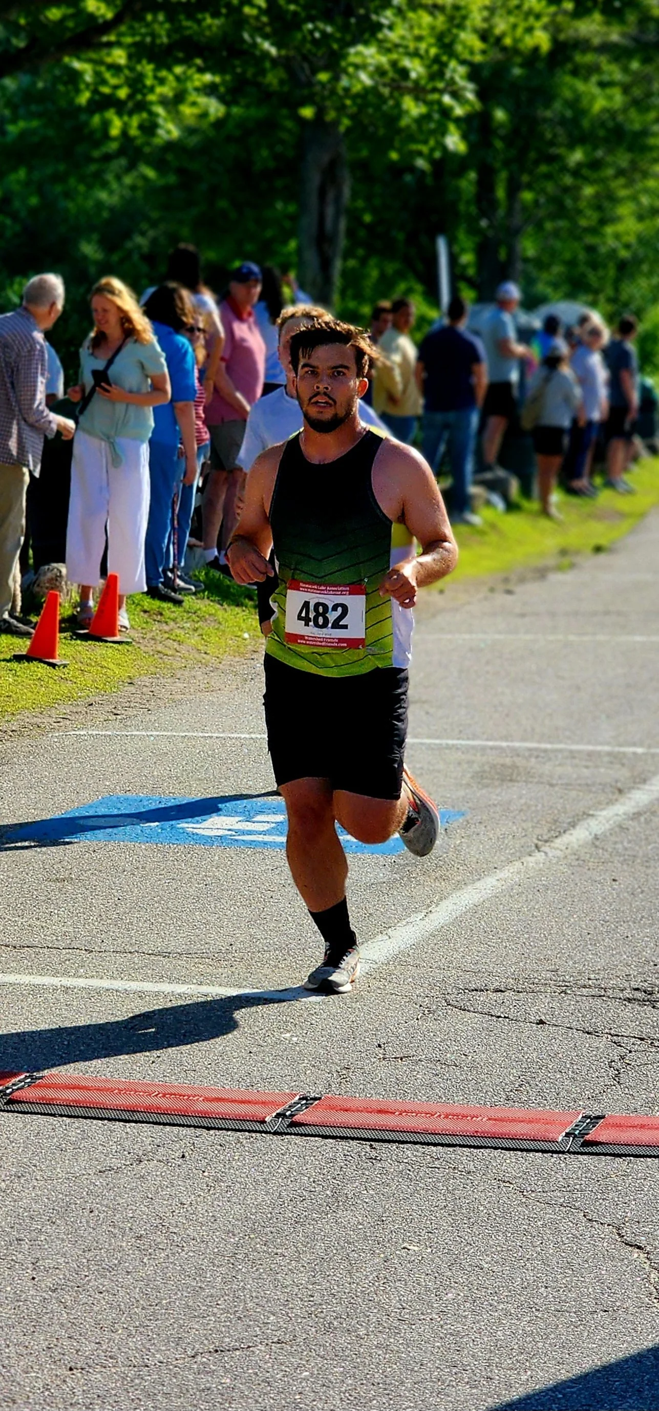 A man wearing a black and green tank top and black shorts running with a crowd standing in the background