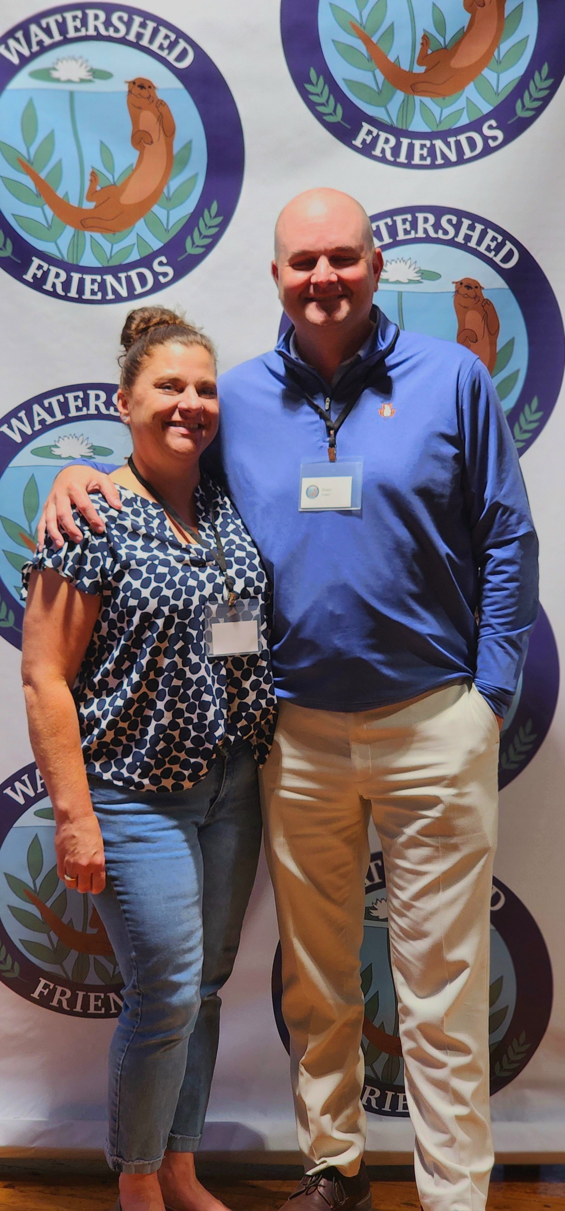 A woman, wearing a blue and white patterned shirt and jeans, and a man, wearing a blue quarter zip and khakis, smiling at the camera