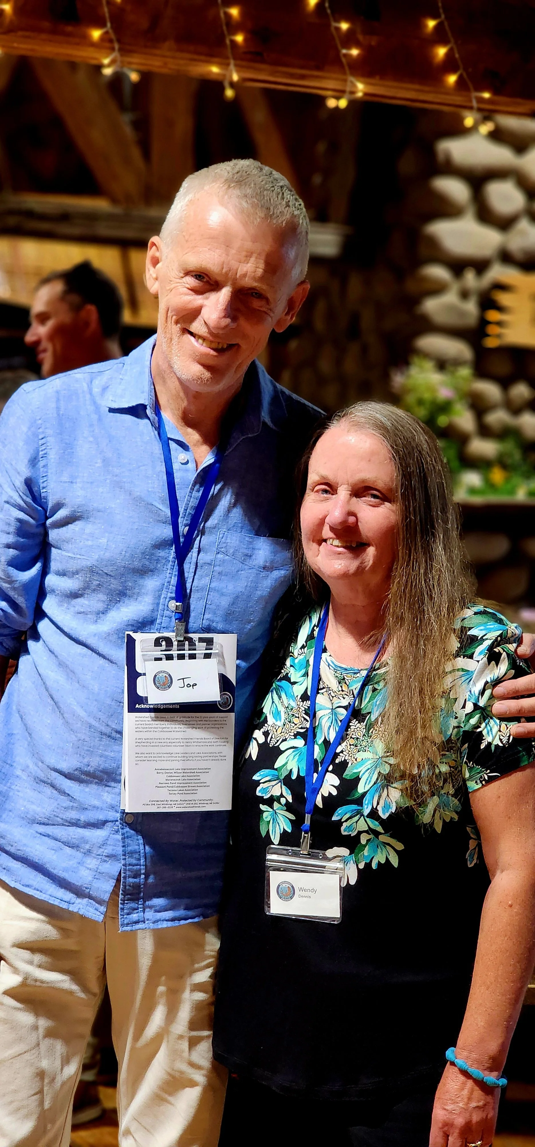 A man, wearing a blue button up shirt and khakis, and a woman wearing a black shirt with a blue and white floral pattern, smiling at the camera