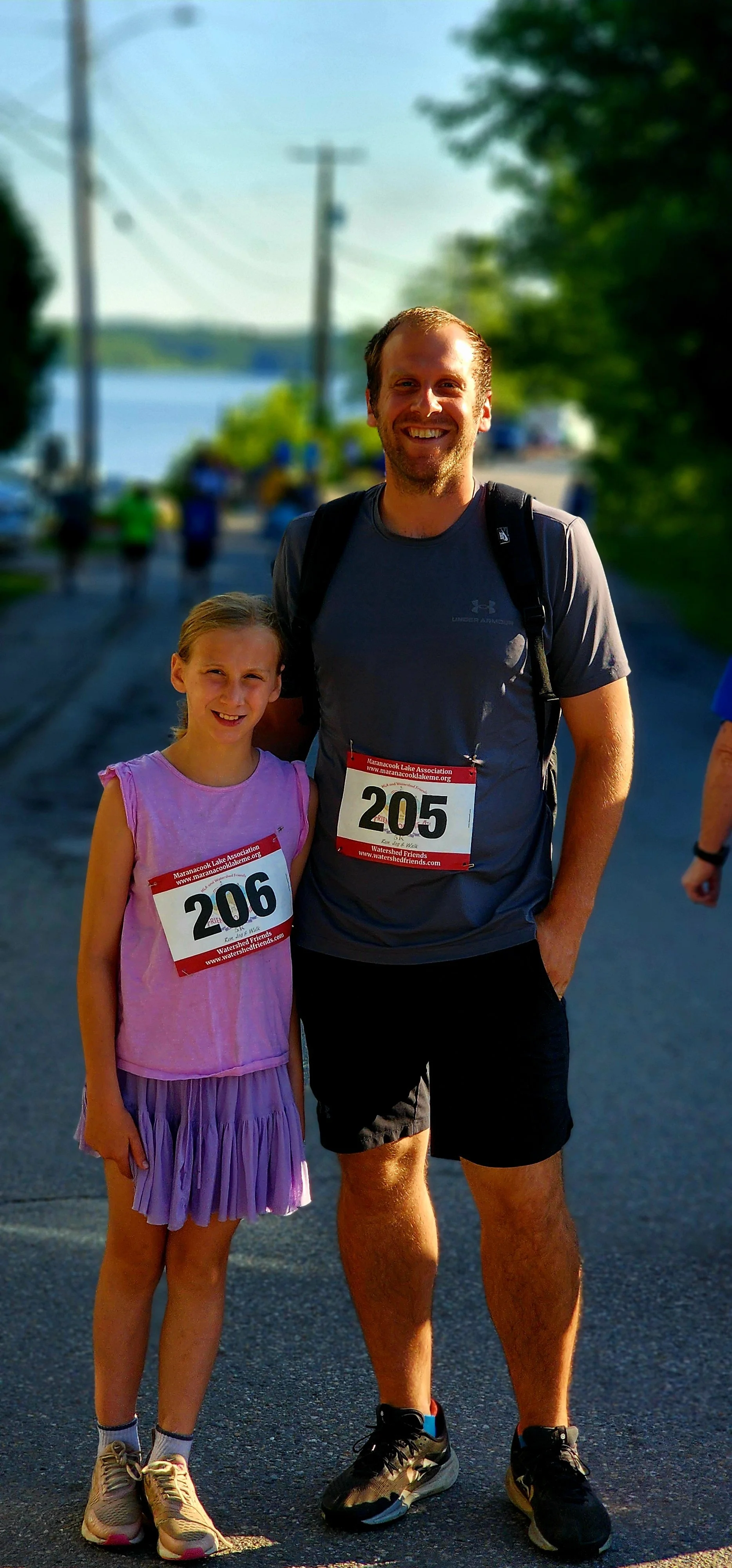 A girl, wearing a a purple tank top and skirt, and a man, wearing a grey t-shirt and black shorts, wearing race bib numbers, smiling at the camera
