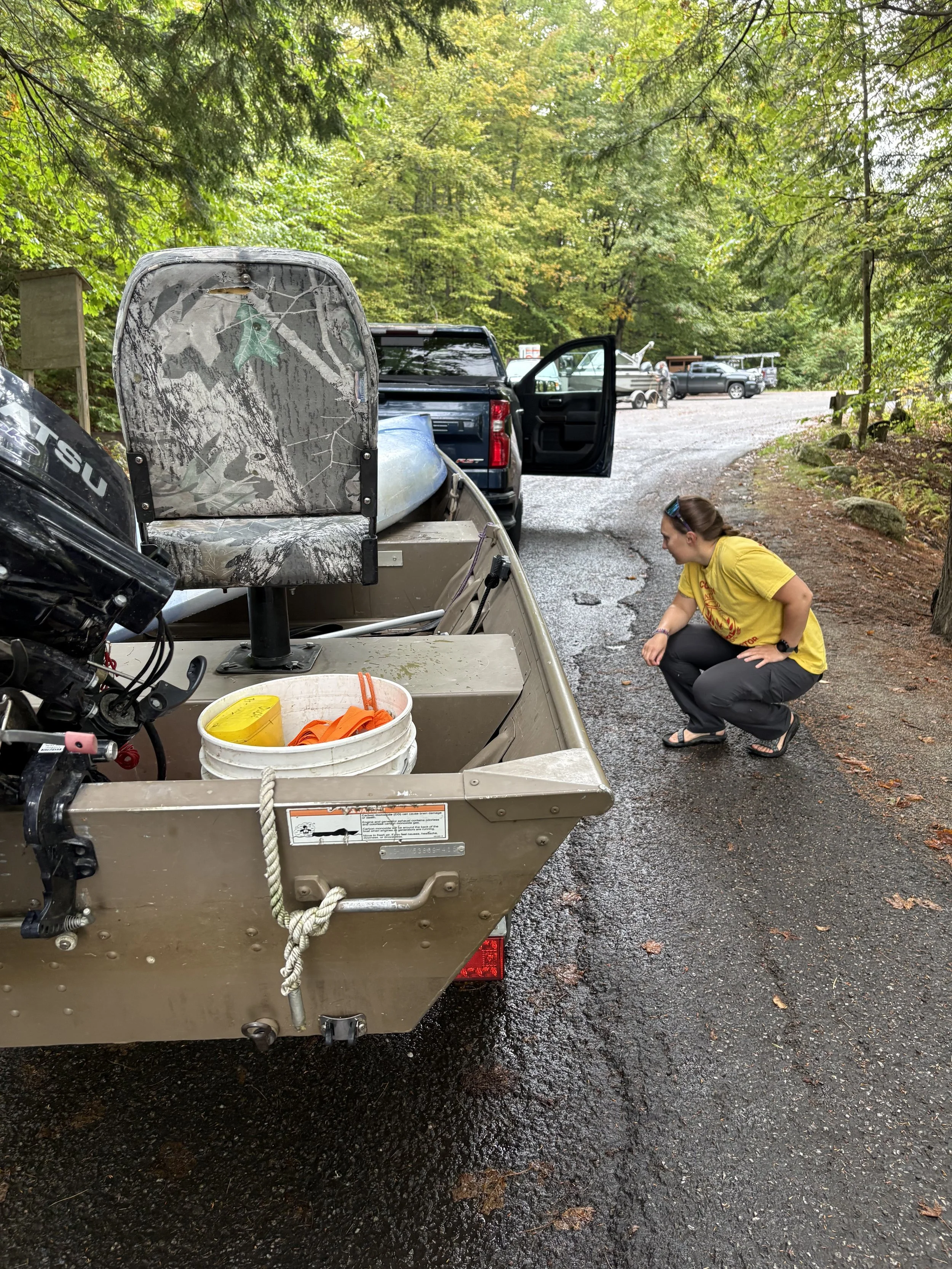 A Friends CBI in a yellow staff shirt crouched beside a small motorboat, looking under the boat at the trailer