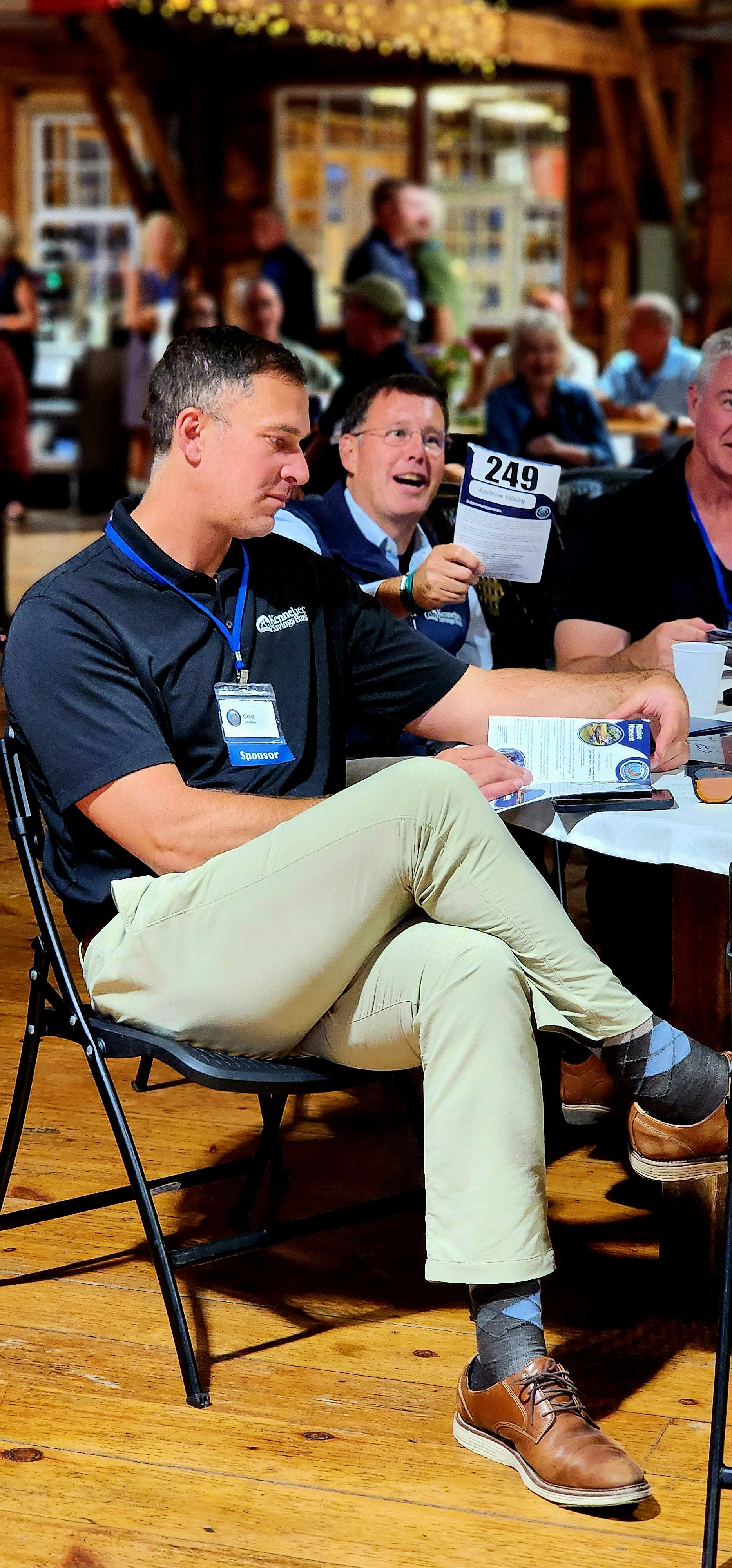 A man, wearing a navy polo with khakis sitting in a chair looking at the event program while another man behind him holds up an event program