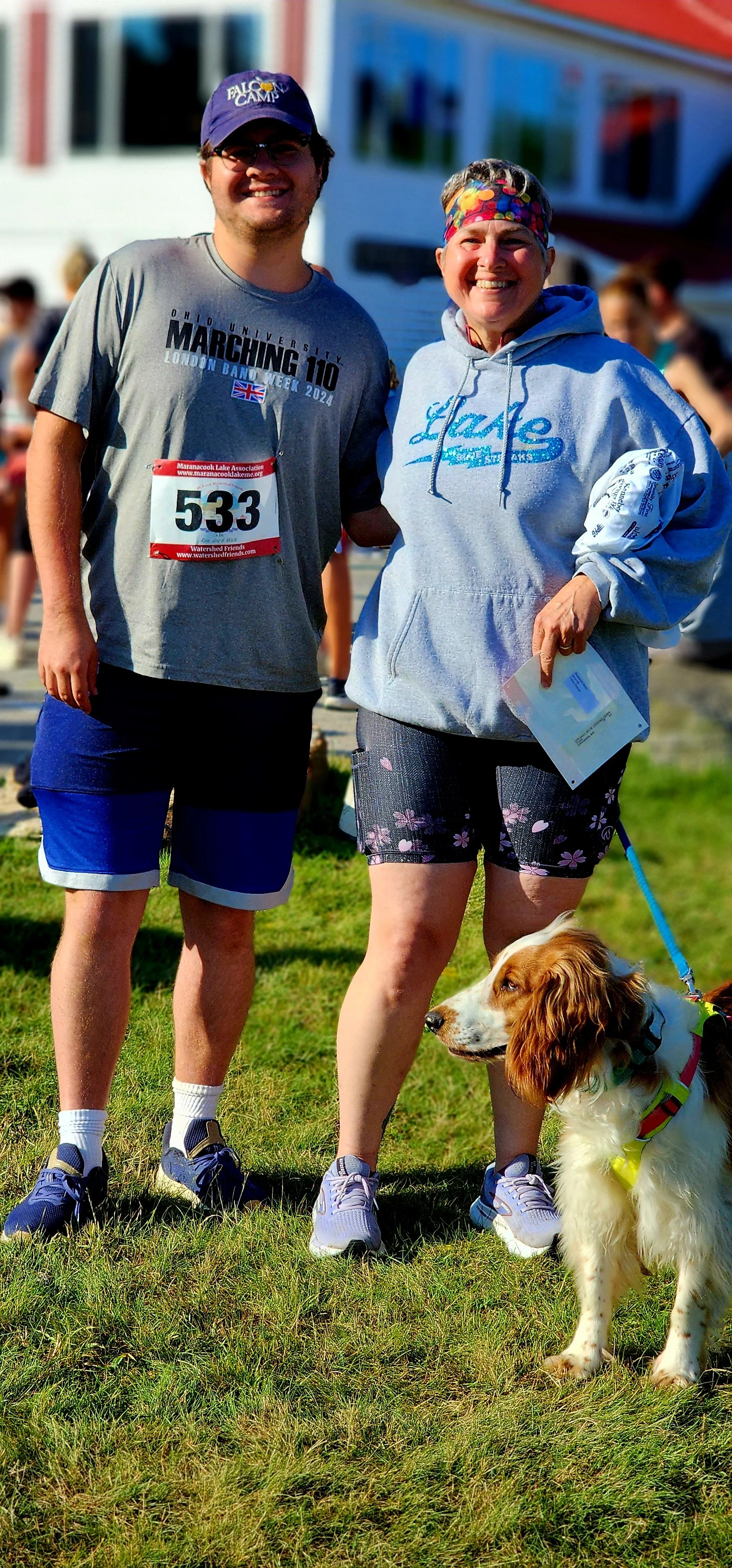 A man, wearing a grey shirt blue shorts and a blue ball cap, and a woman, wearing a grey sweatshirt and black shorts, smiling at the camera with a dog
