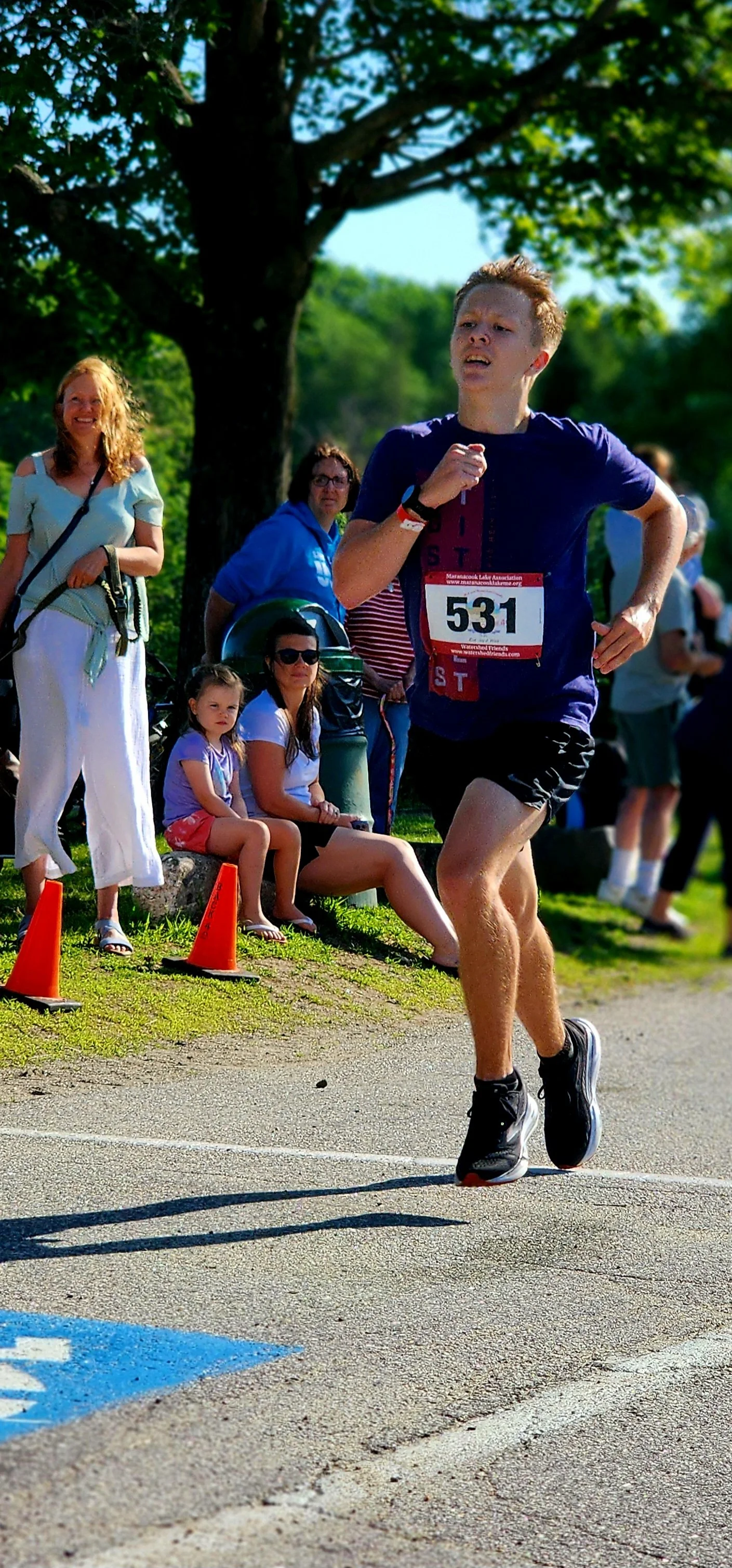 A man wearing a dark blue t-shirt and black shorts running with a crowd standing behind him