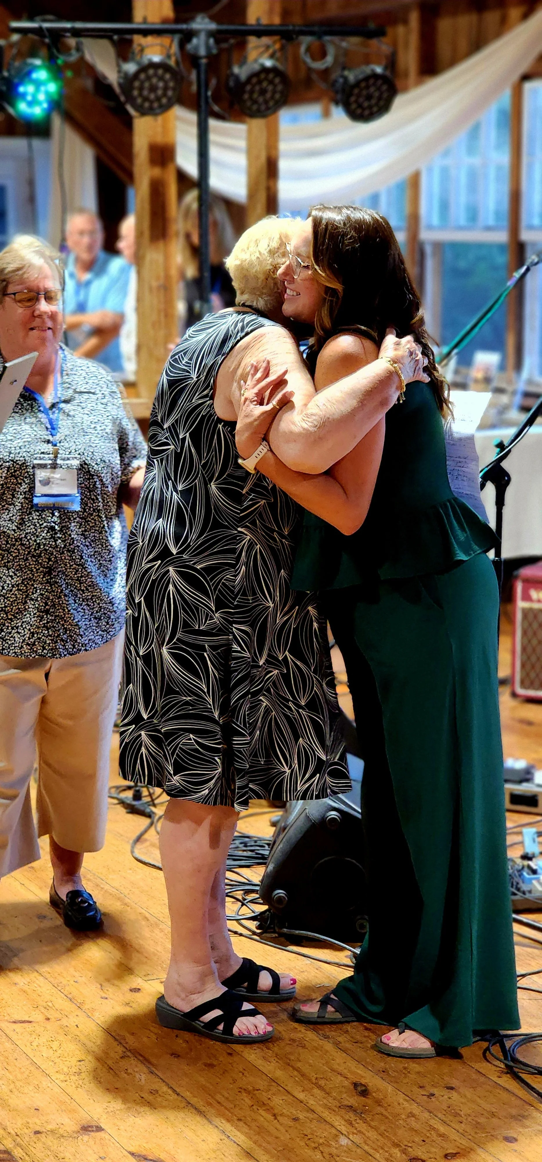 Two women, one wearing a black dress with white patterns the other wearing a green jumpsuit, hugging