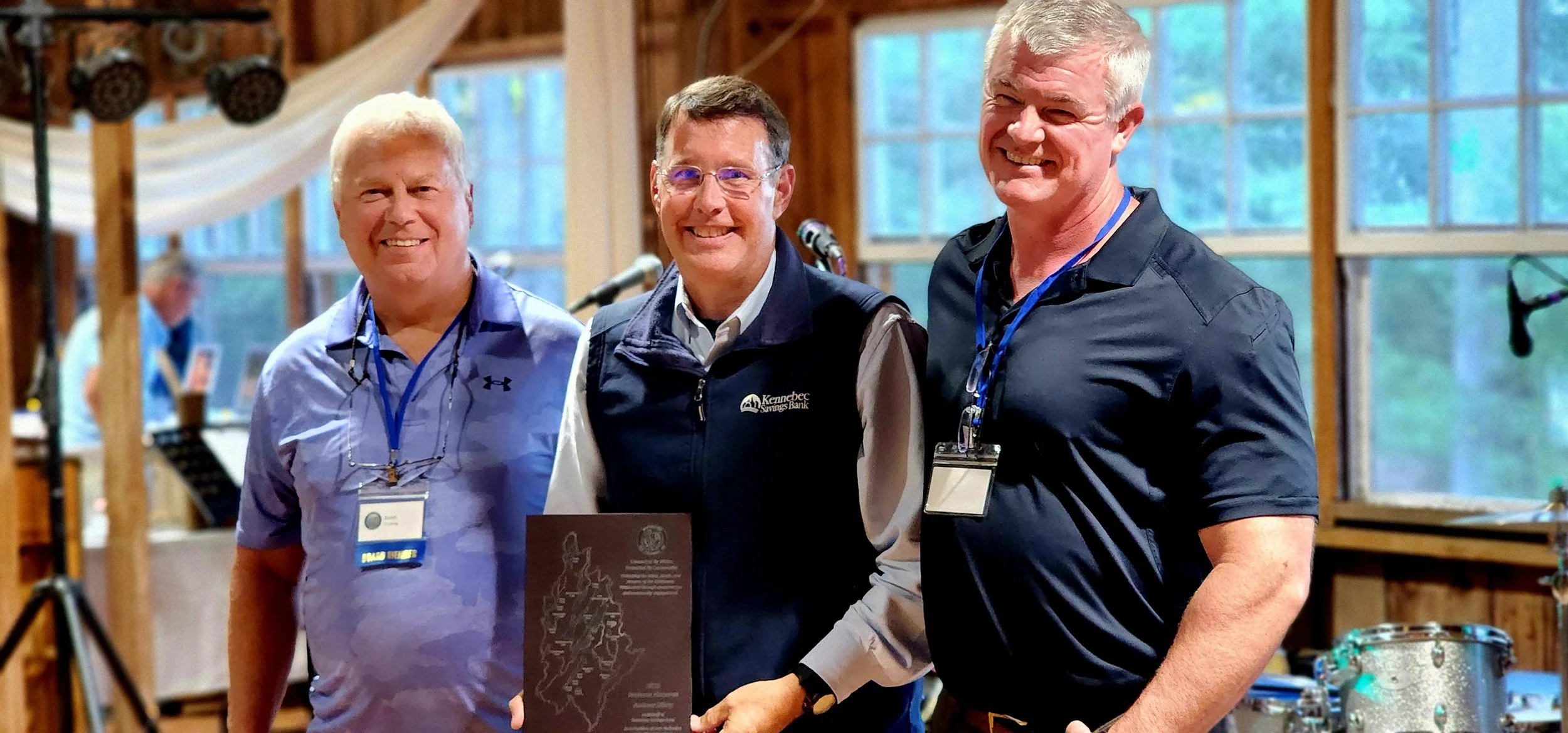 Three men smiling at the camera, the man in the middle is holding a plaque