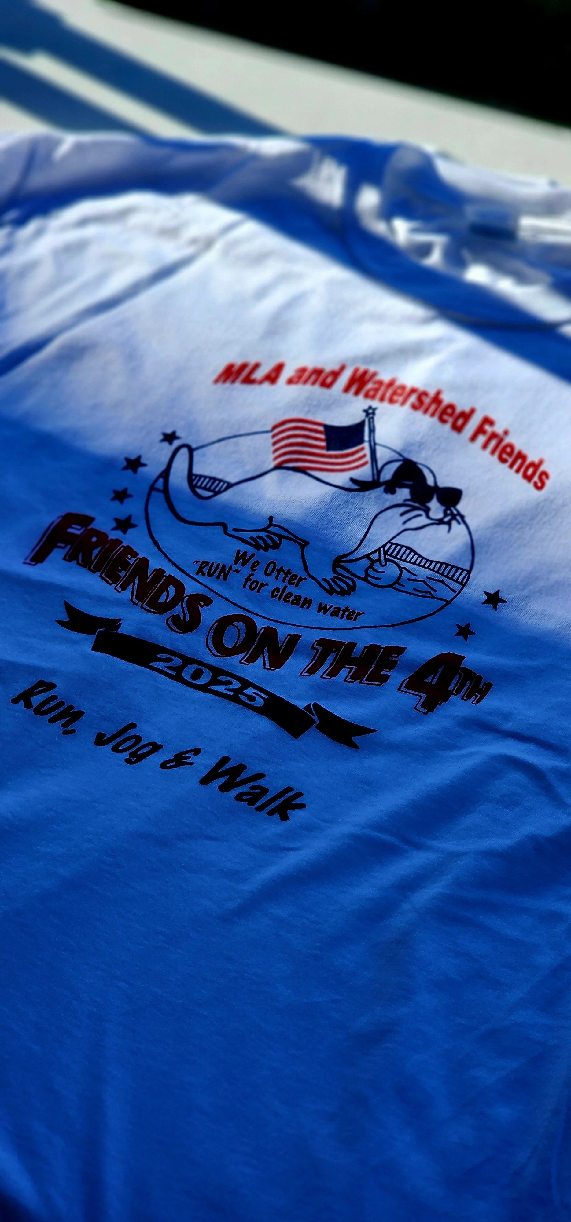 The front of the race t-shirt, showing the race logo which has a cartoon otter wearing sunglasses and a backwards hat carrying an American flag, along with the words "MLA and Watershed Friends, Friends of the 4th 2025, run, jog, and walk"