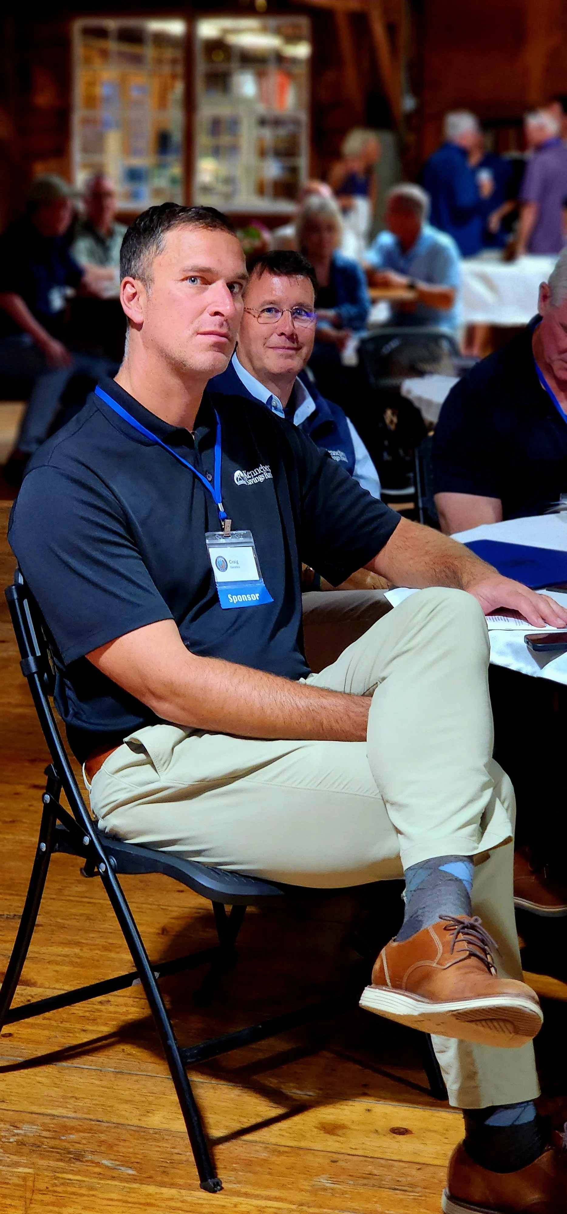 A man, wearing a navy polo and khakis, sitting in a chair watching something off camera
