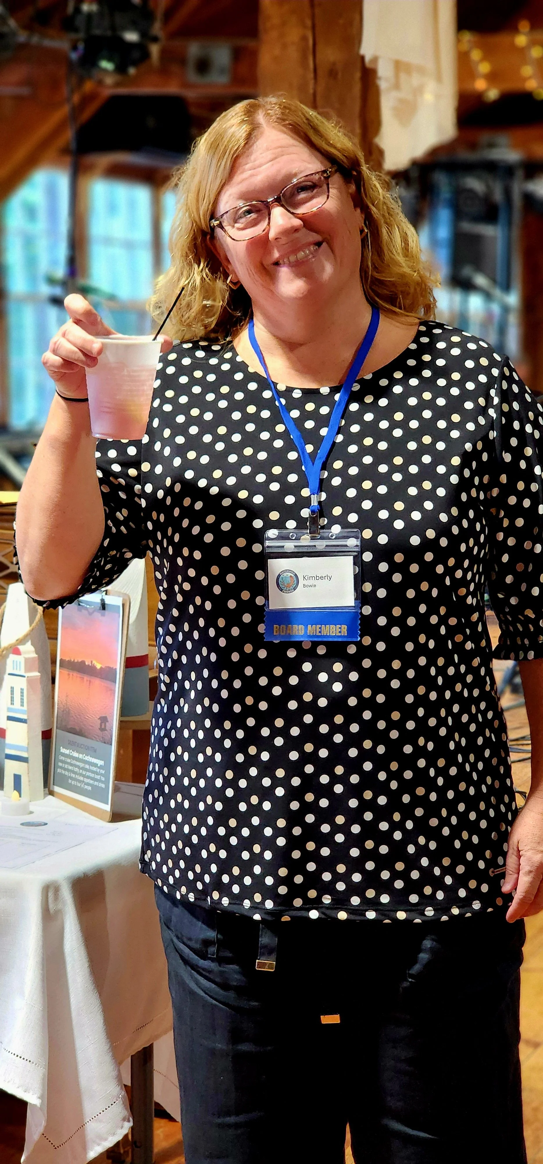 A woman, wearing a black shirt with white polka dots and black pants, smiling at the camera, holding up a plastic cup with a beverage in it