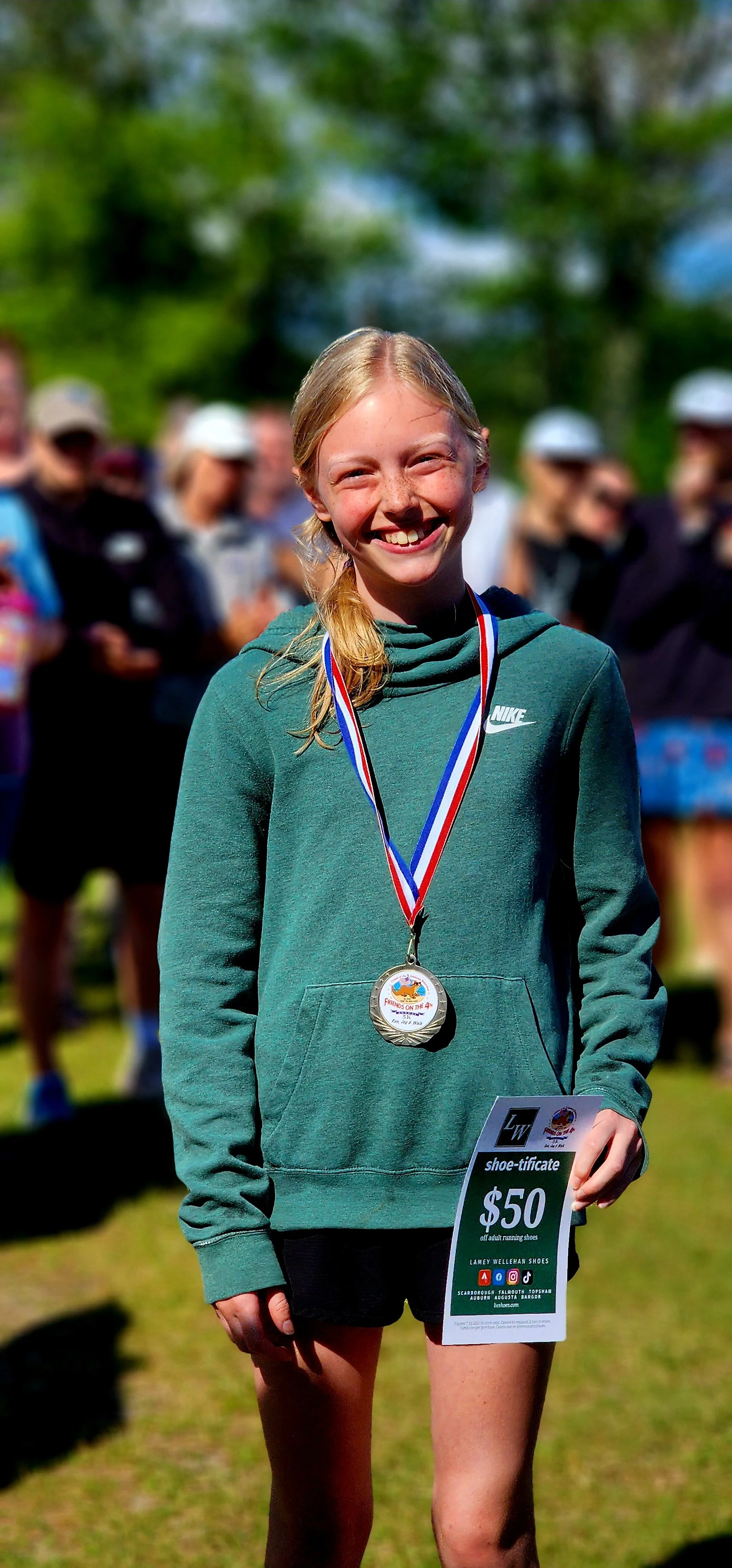 A girl smiling at the camera, wearing an award medal, green sweatshirt, black shorts and holding a certificate to Lamey Wellehan shoes