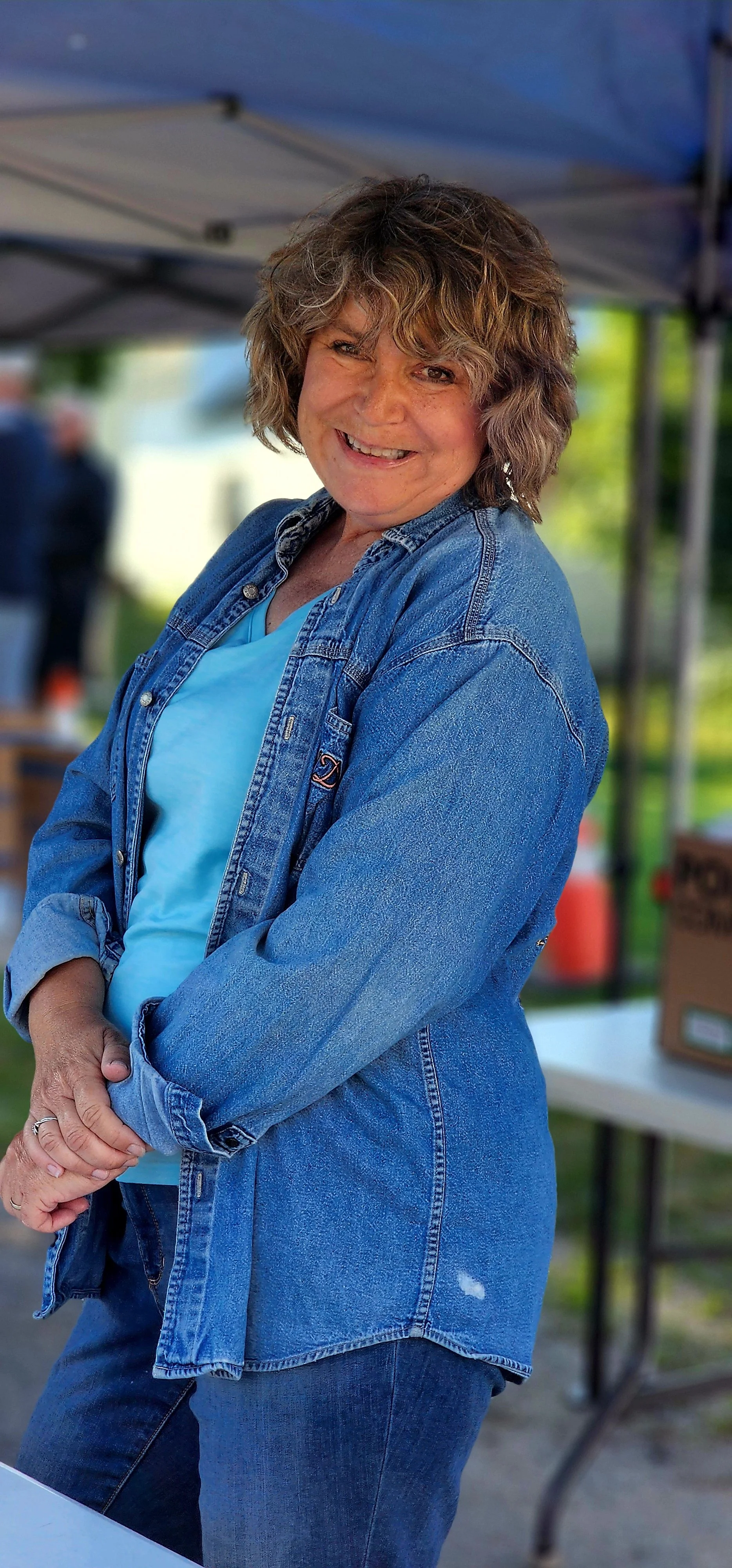 A woman, wearing a jean jacket over a blue shirt and jeans, smiling at the camera