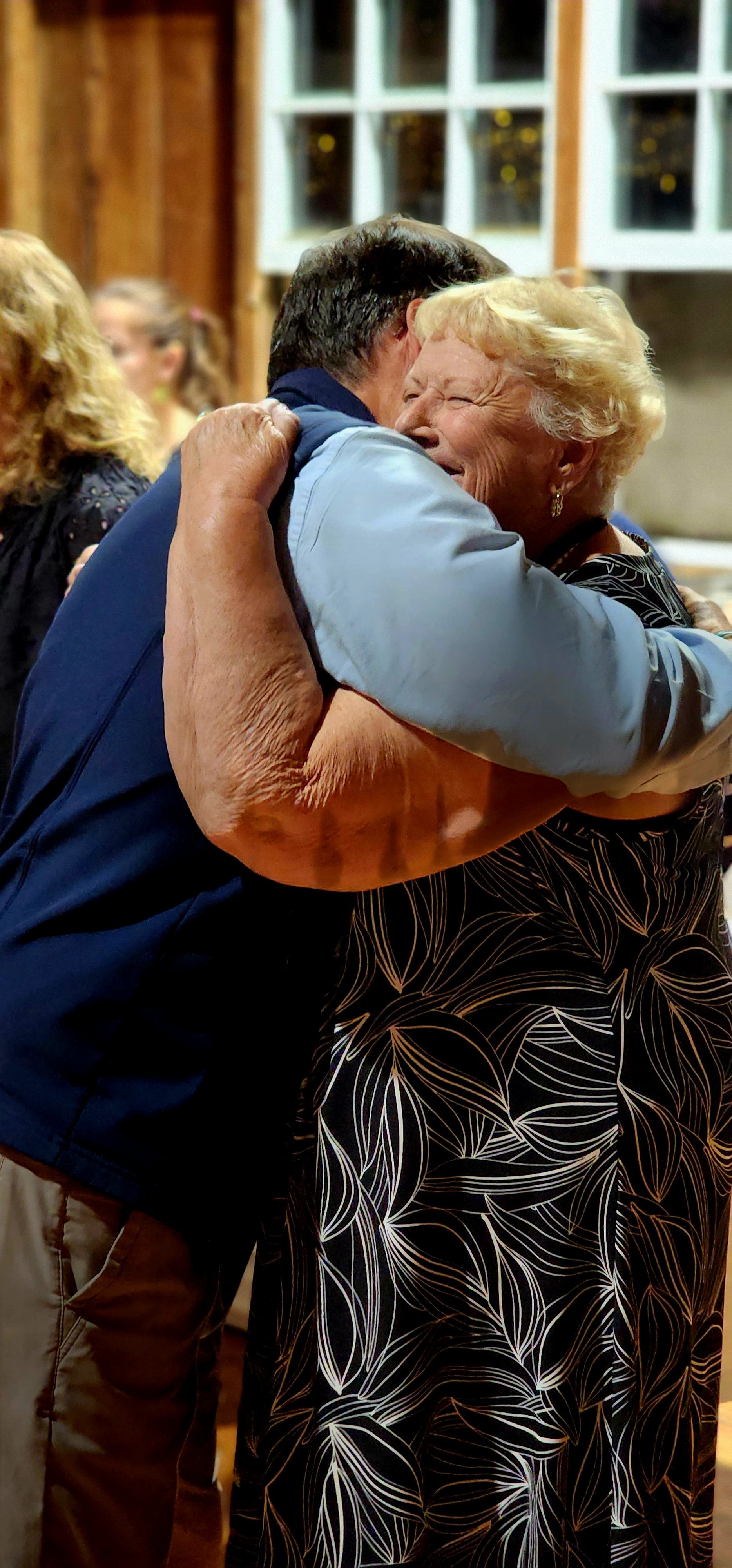 A man, wearing a dark blue vest over a light blue long sleeve and khakis, and a woman, wearing a black dress with white patters, hugging