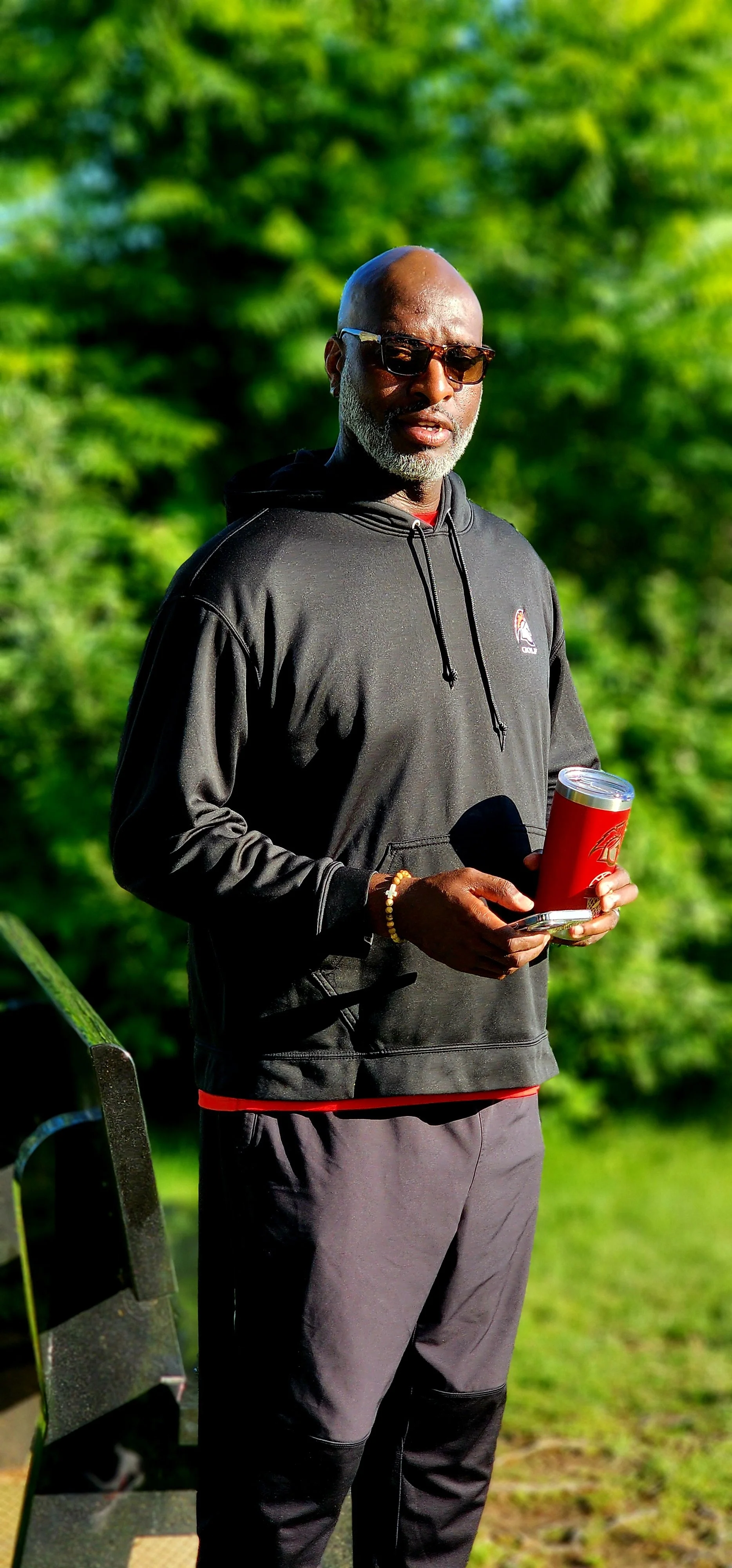 A man, wearing a grey sweatshirt and grey pants, holding a travel mug and smiling at the camera
