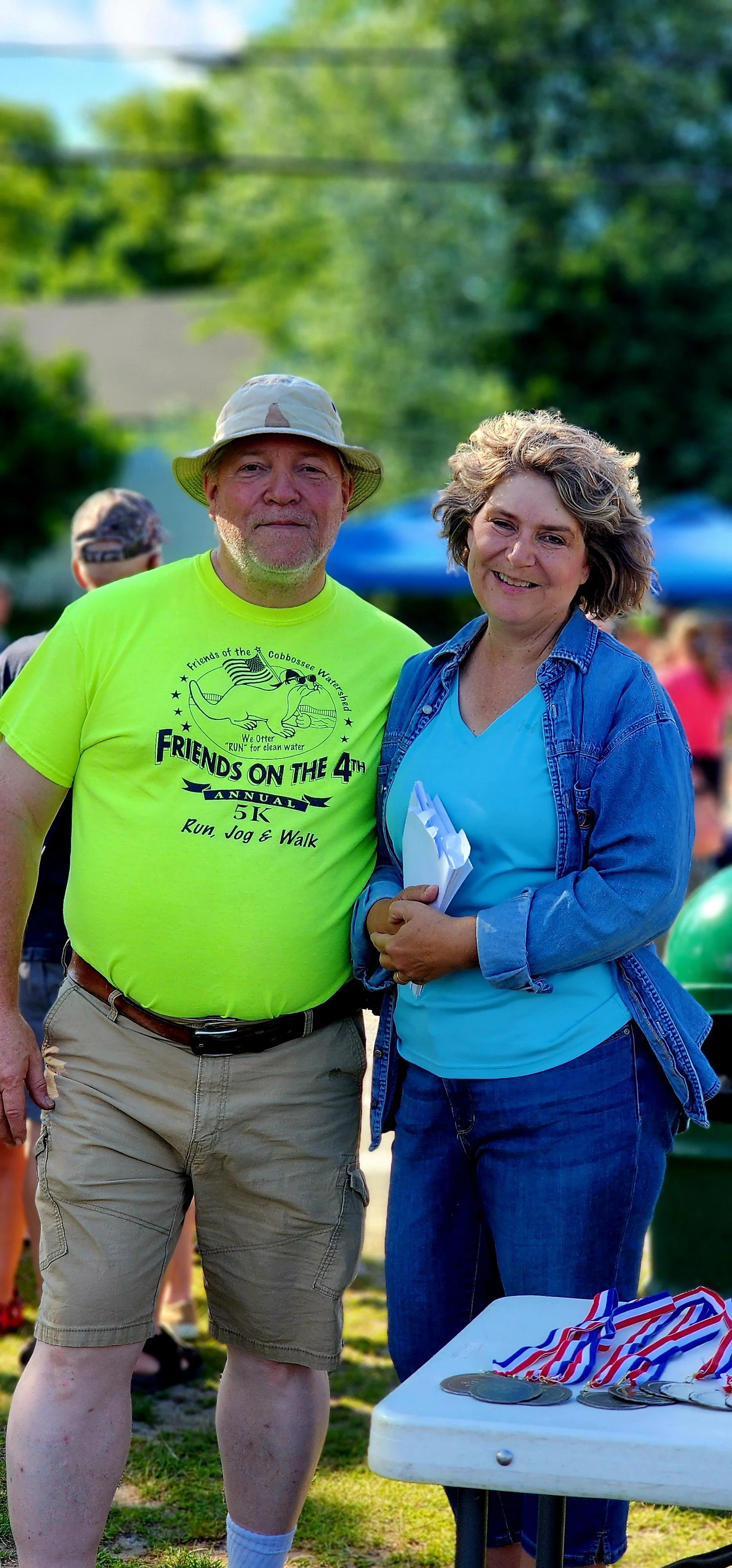 A man wearing a neon yellow t-shirt and khaki shorts standing with a woman wearing a jean jacket over a blue shirt and jeans