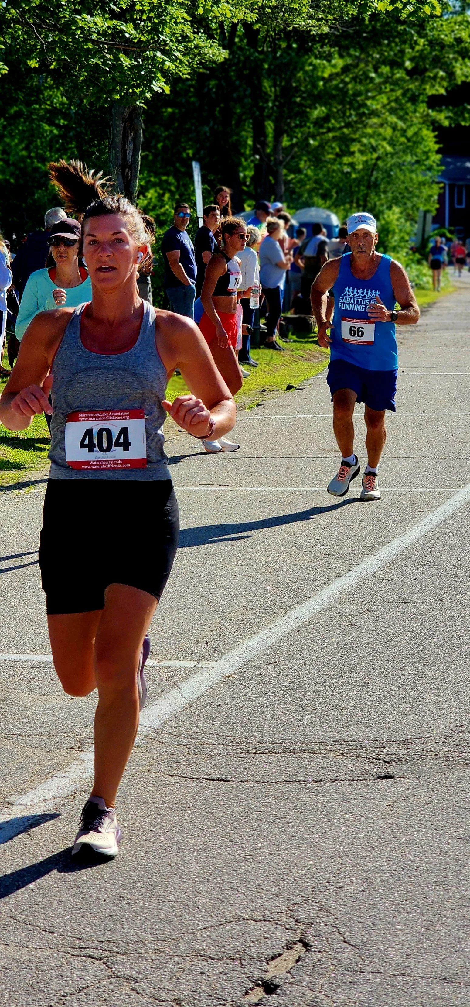 A woman wearing a grey tank top and black shorts running ahead of a man wearing a blue tank top and black shorts with a crowd standing in the background