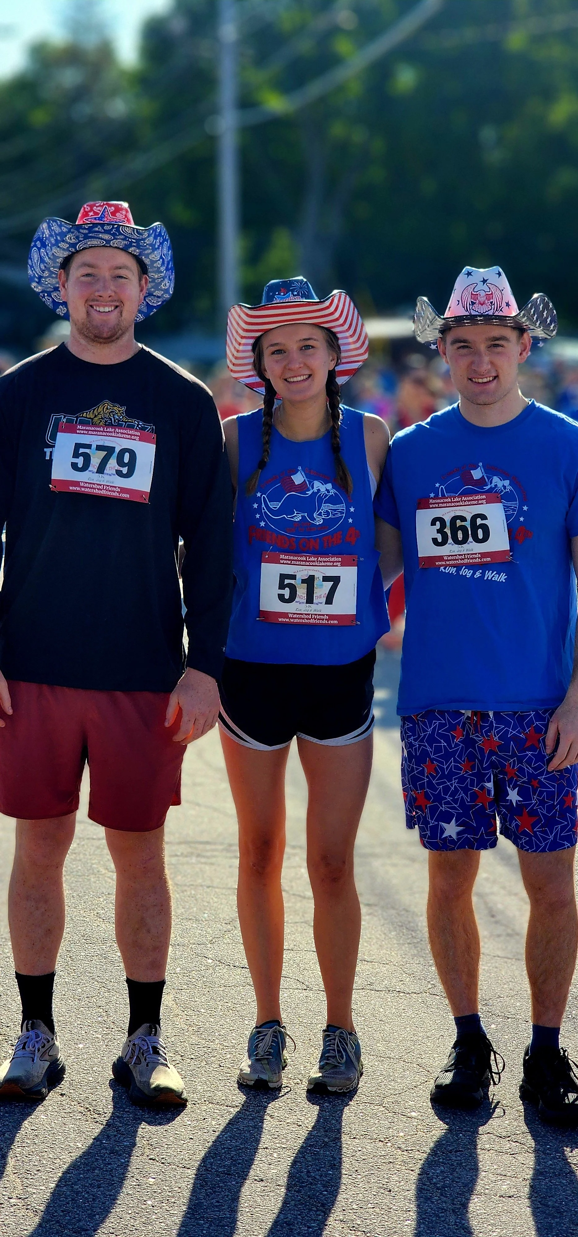 Three people wearing patriotic cowboy hats, one man wearing a black long sleeve shirt and red short, a  woman wearing a blue t-shirt and black shorts, and a man wearing a blue t-shirt and blue shorts, smiling at the camera