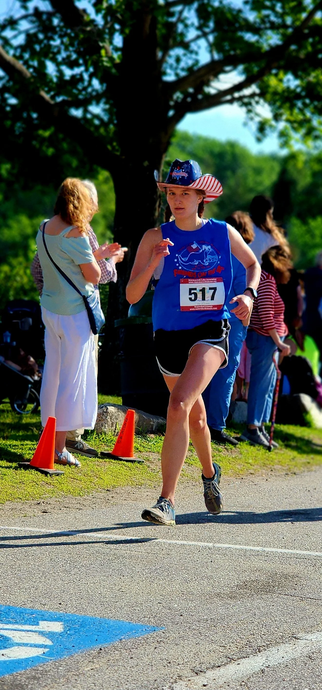 A person wearing a patriotic cowboy hat, blue t-shirt, and black shorts running with a crowd standing in the background