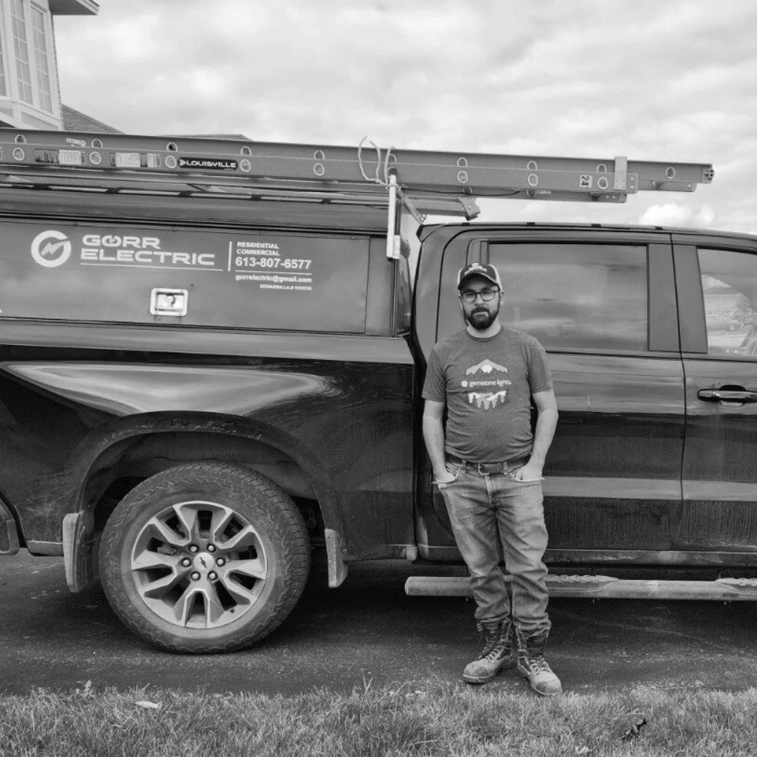 A man standing next to a black pickup truck with a ladder on top. The truck has a sign for Gorbe Electric and an email address. The man is wearing a cap, glasses, a T-shirt, jeans, and work boots, and is standing on a grassy area.