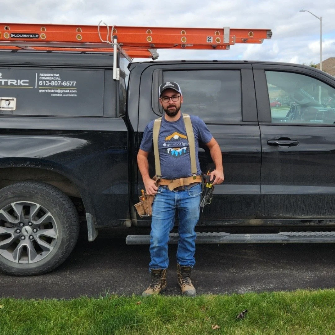 A man in work gear standing in front of a black utility truck with a ladder on top, on a street with grass in the foreground and a cloudy sky above.