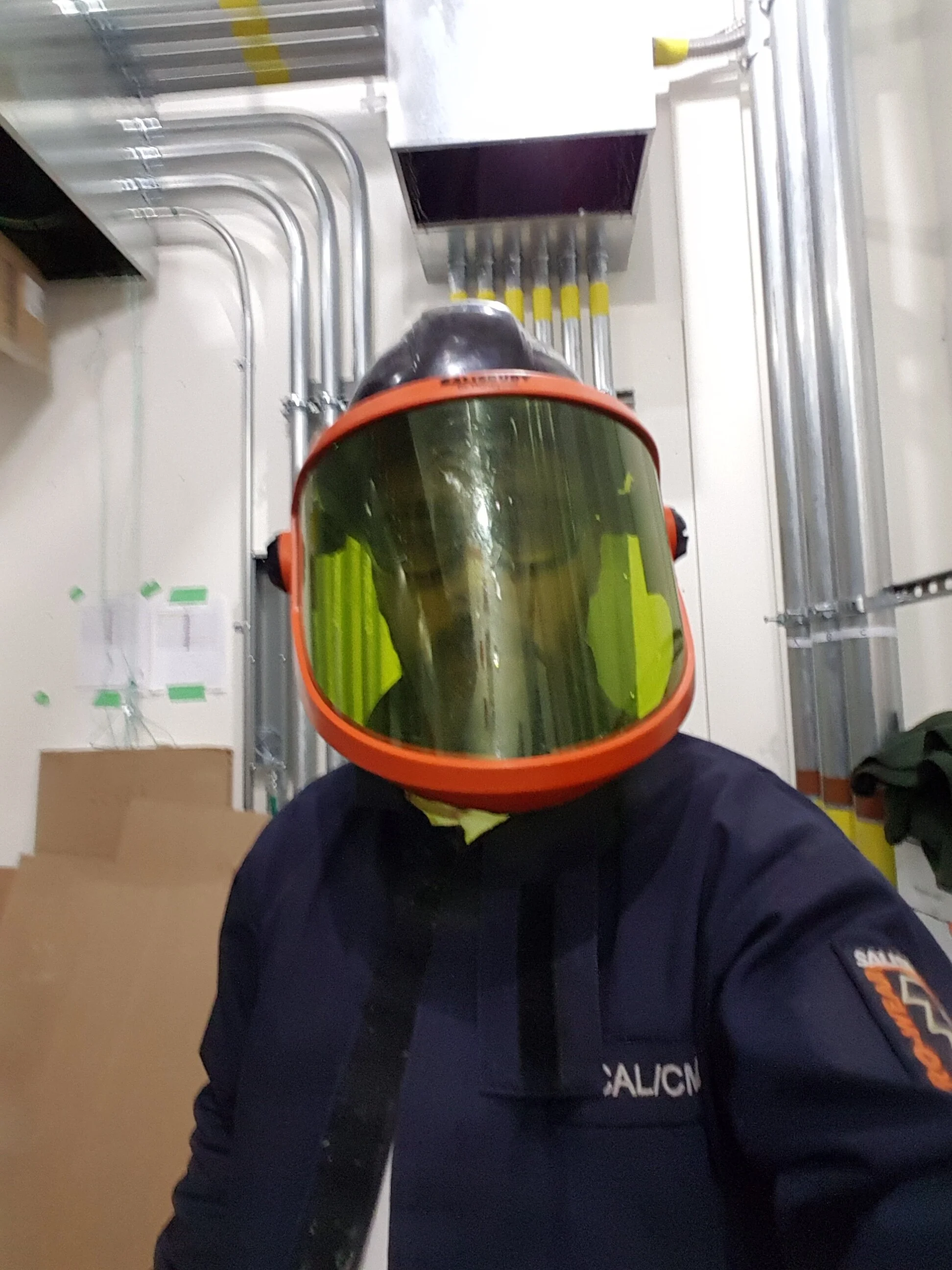 Person wearing a hazmat suit and mask taking a selfie in an industrial room with pipes and boxes.