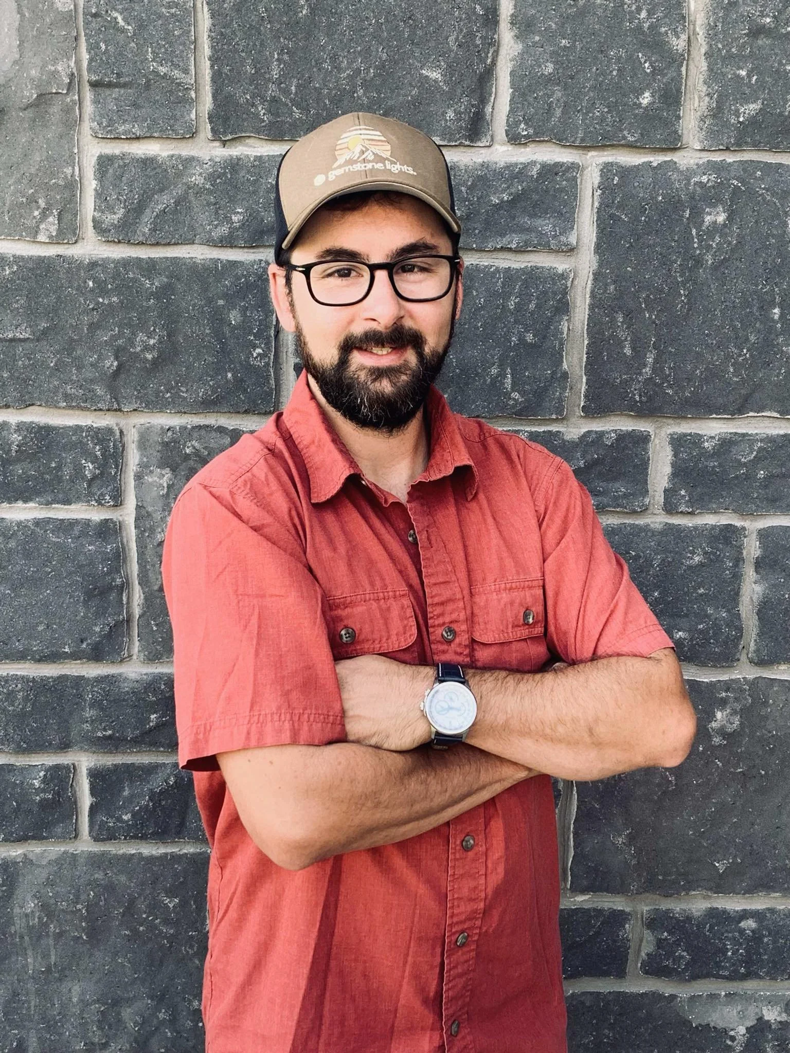 A man with glasses and a beard, wearing a red short-sleeved button-up shirt and a cap, standing with arms crossed in front of a gray brick wall.