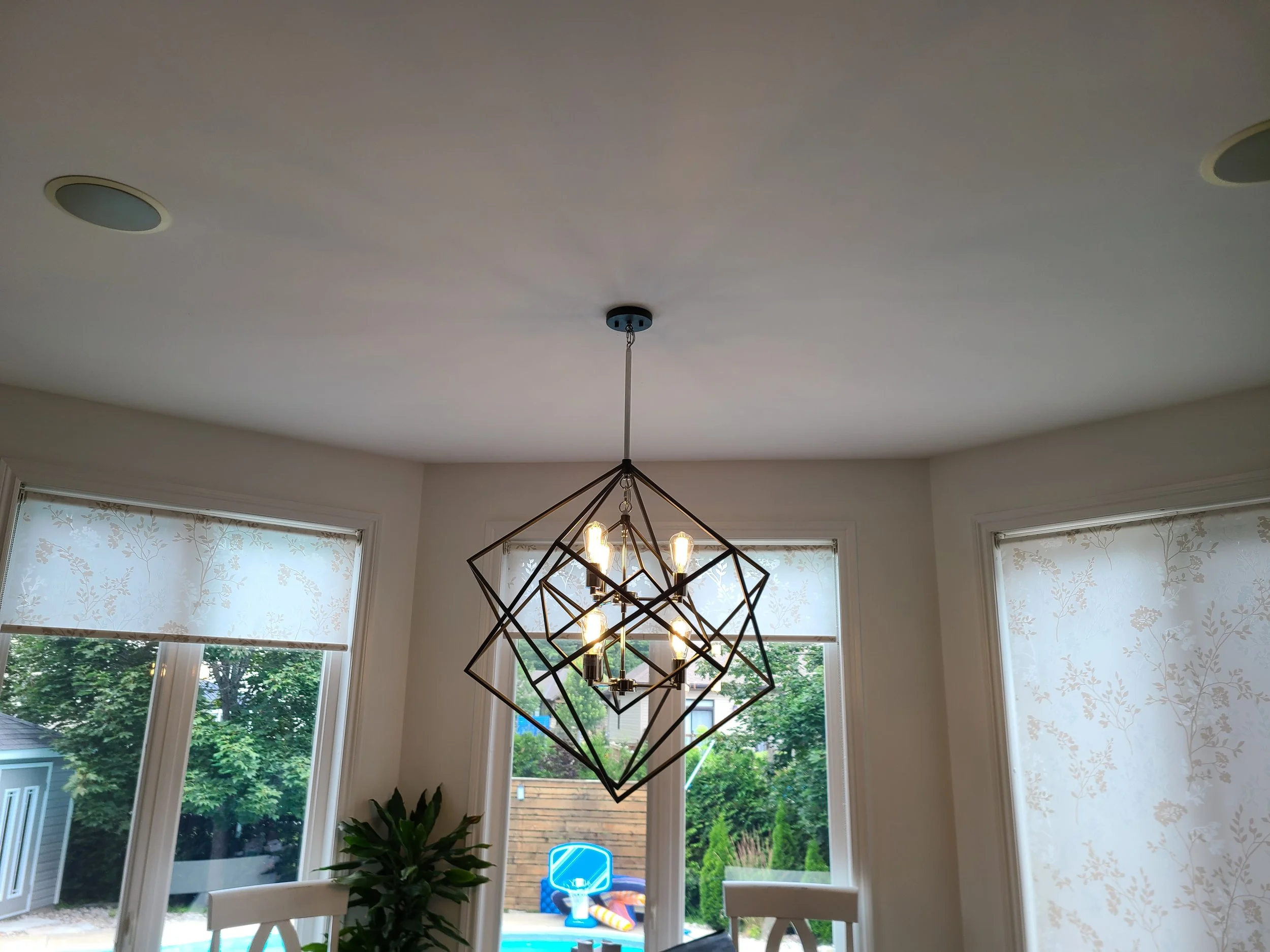 Modern geometric chandelier hanging from a white ceiling, with large windows showing greenery and a backyard patio with a blue and yellow trampoline