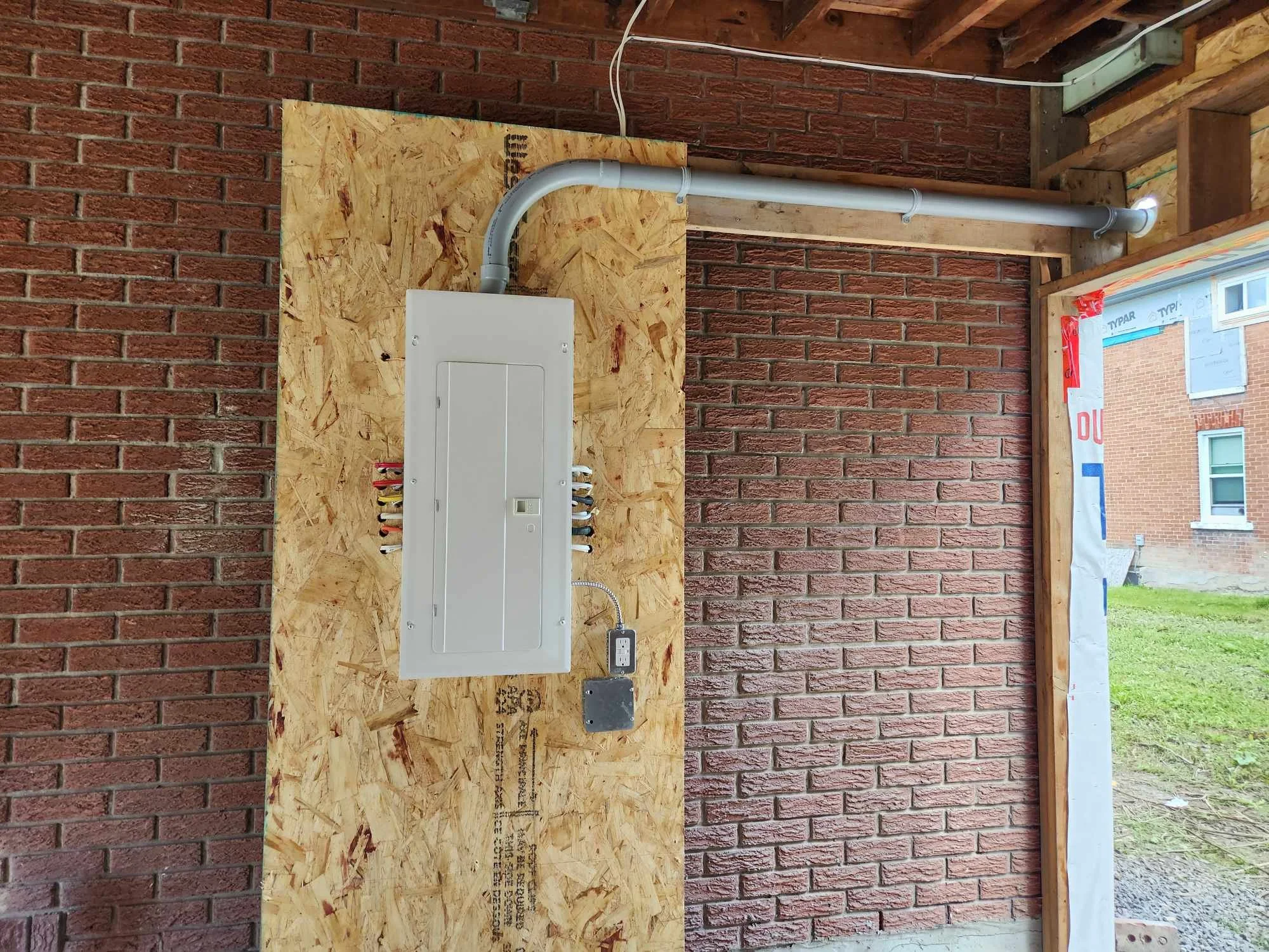 Interior view of a utility box mounted on an OSB board with a metal conduit pipe running to the right wall of a brick structure under construction.