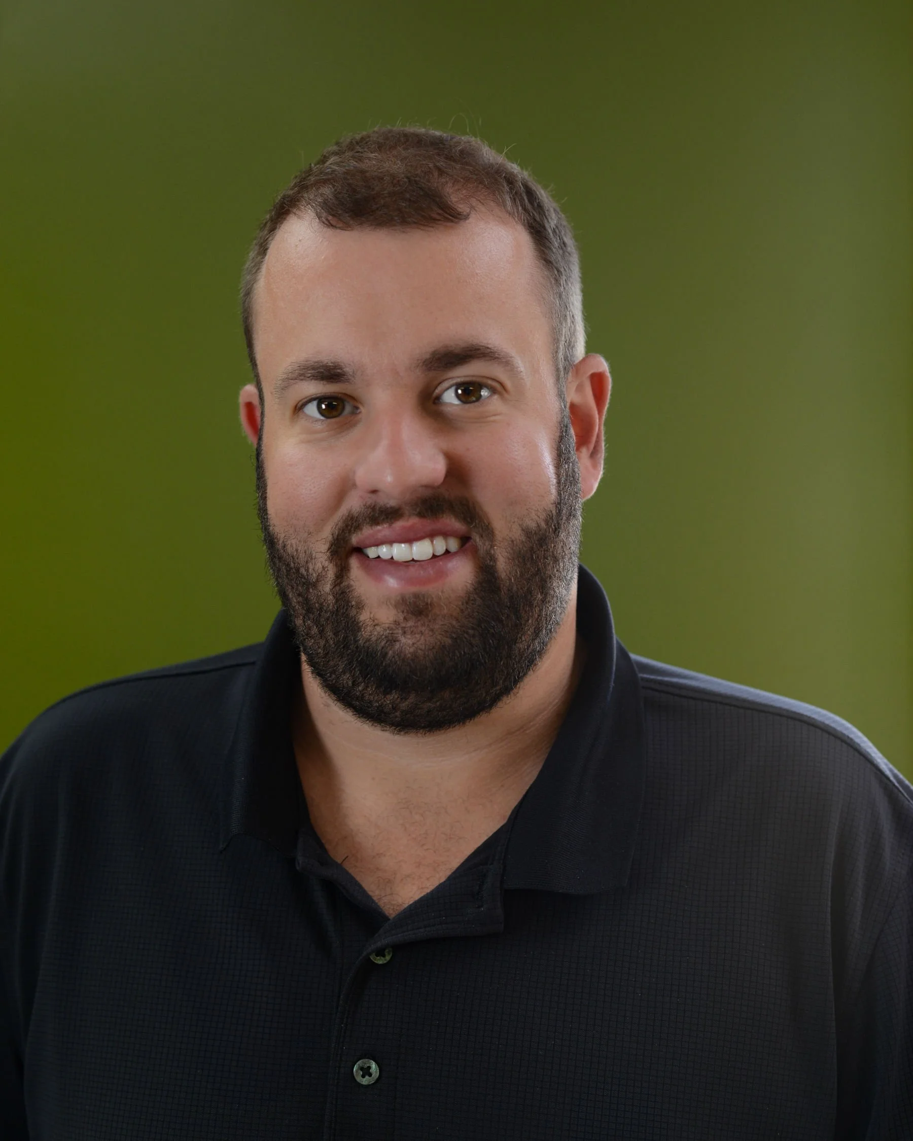 Close-up of a smiling man with brown hair and beard wearing a black shirt against a green background.