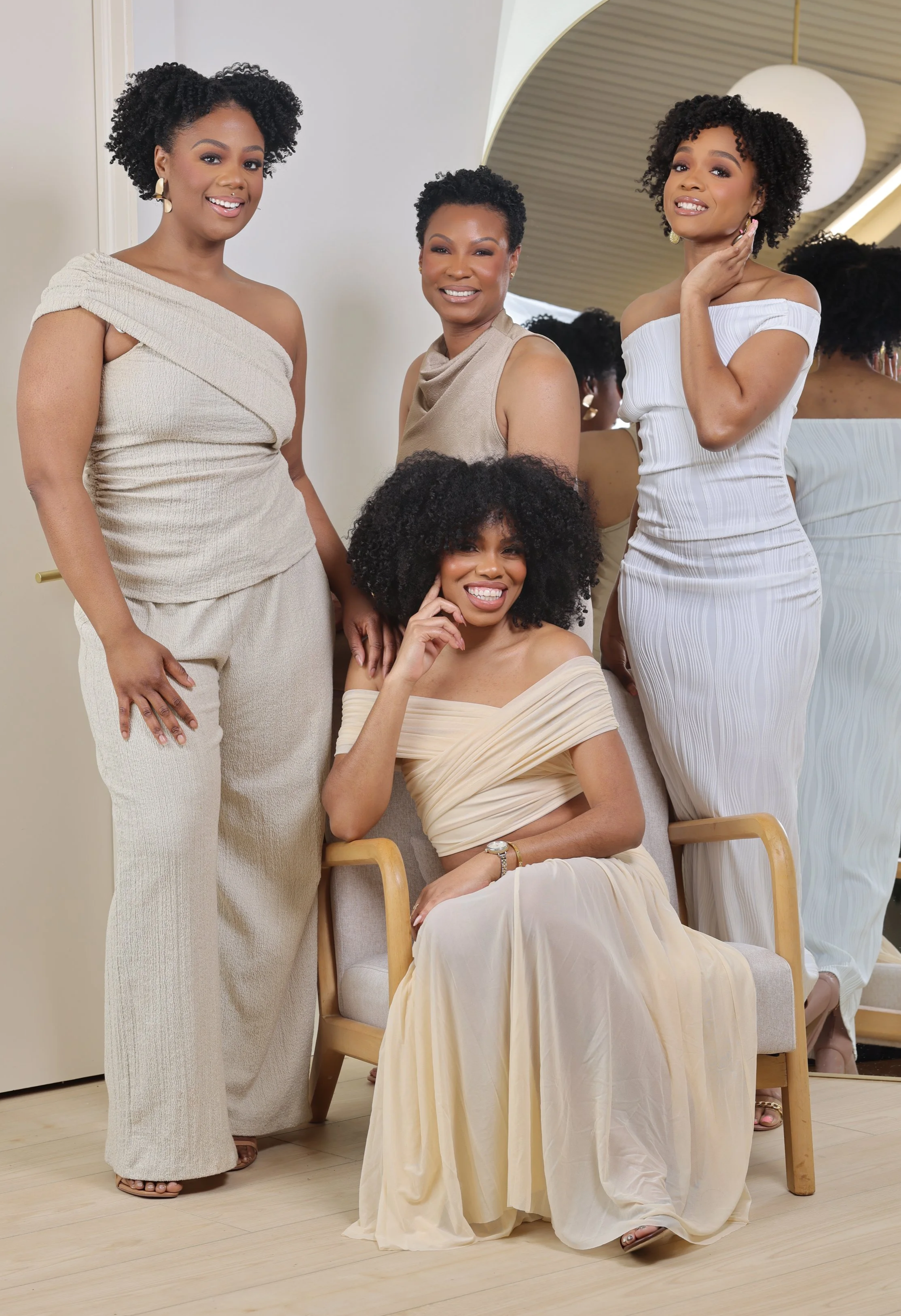 Four women with curly hair in elegant beige and white dresses, posing together in front of a mirror in a well-lit room.