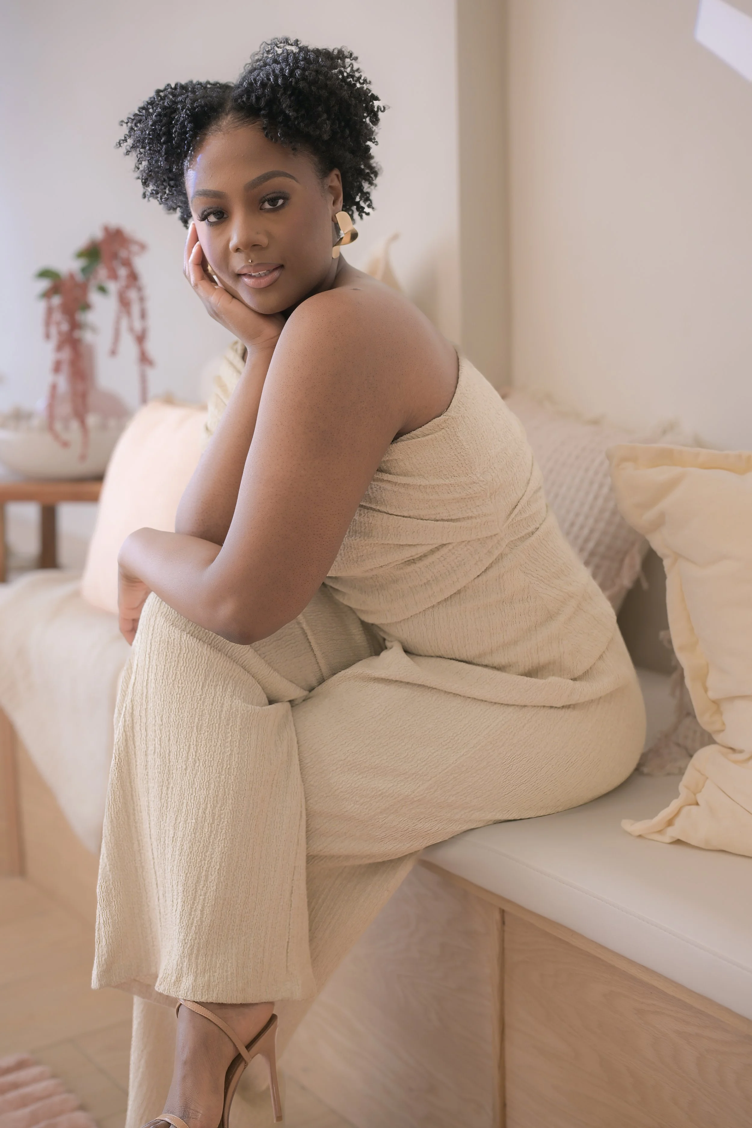 A woman sitting on a bed in a beige outfit, posing with her hand on her face, in a softly lit bedroom.