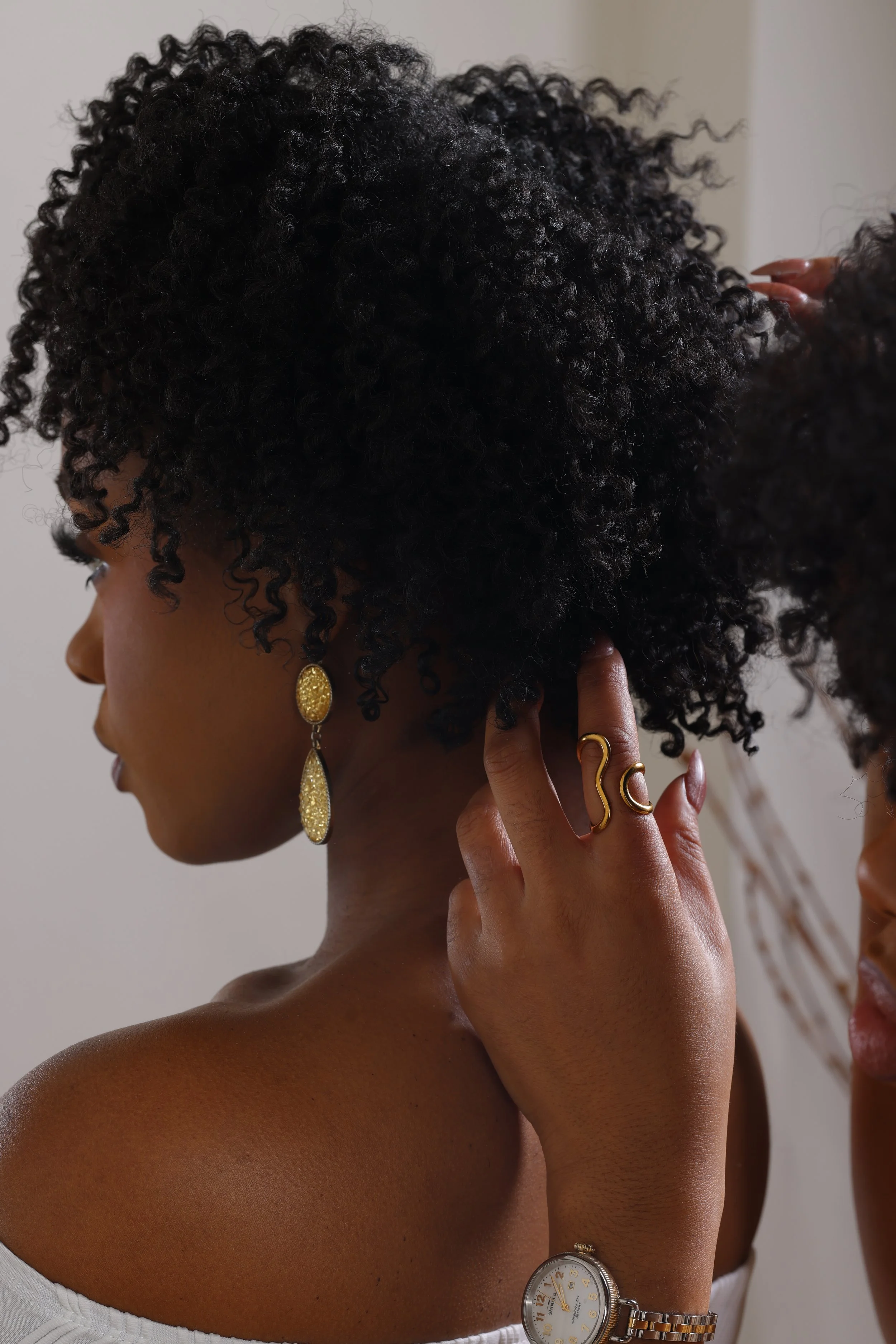 A woman with curly black hair wearing gold earrings, gold rings, and a wristwatch, touching her ear, with part of another person's face visible.