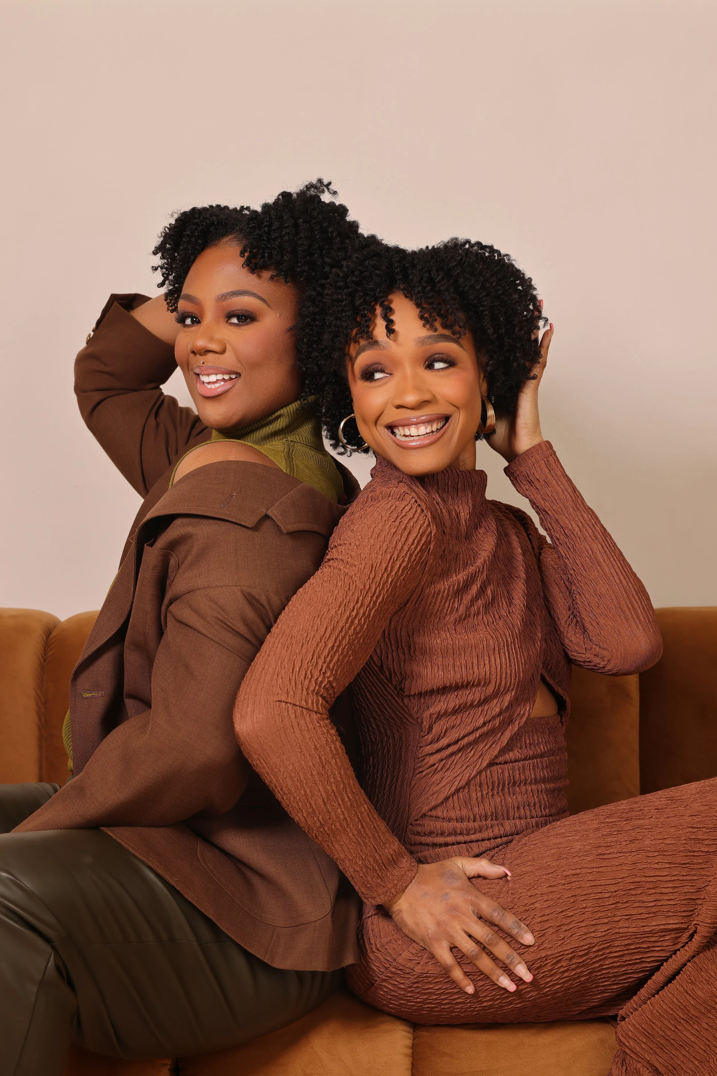 Two smiling women with curly hair sitting on a tan sofa, back to back, facing opposite directions, in a cozy indoor setting.