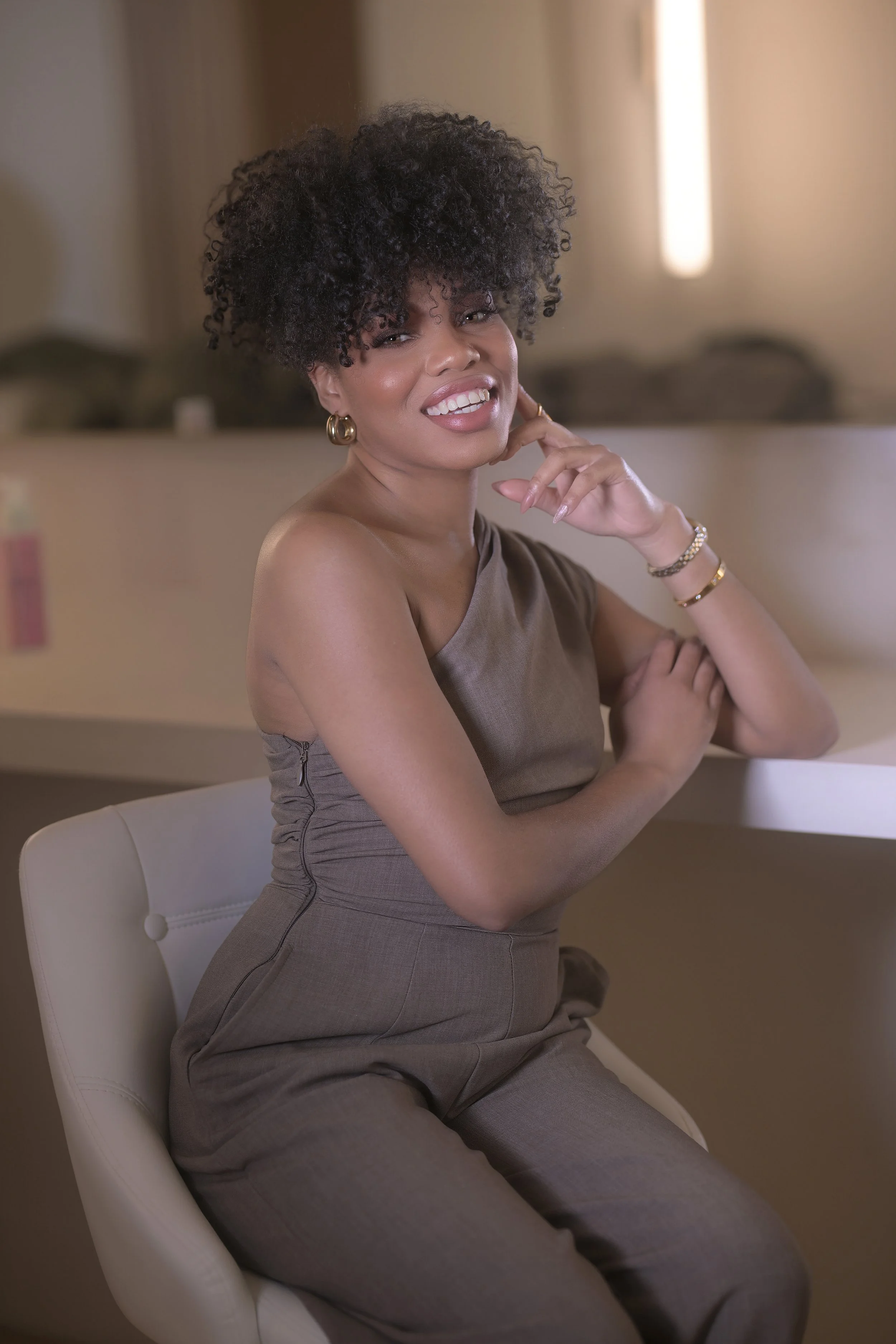 A woman with short curly hair, wearing gold hoops, a brown sleeveless top, and matching pants, sitting on a white chair in a warmly lit room, smiling and looking at the camera.