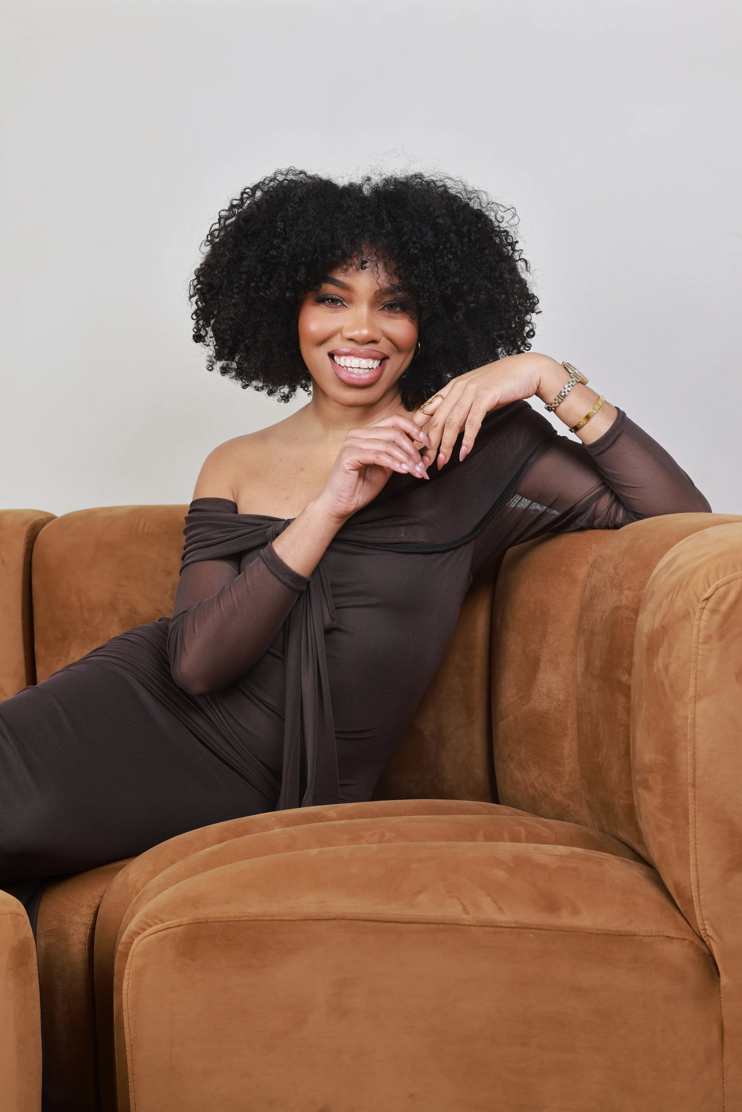 A smiling woman with curly black hair sitting on a brown couch, wearing an off-the-shoulder black dress and jewelry, while leaning comfortably on the couch armrest.
