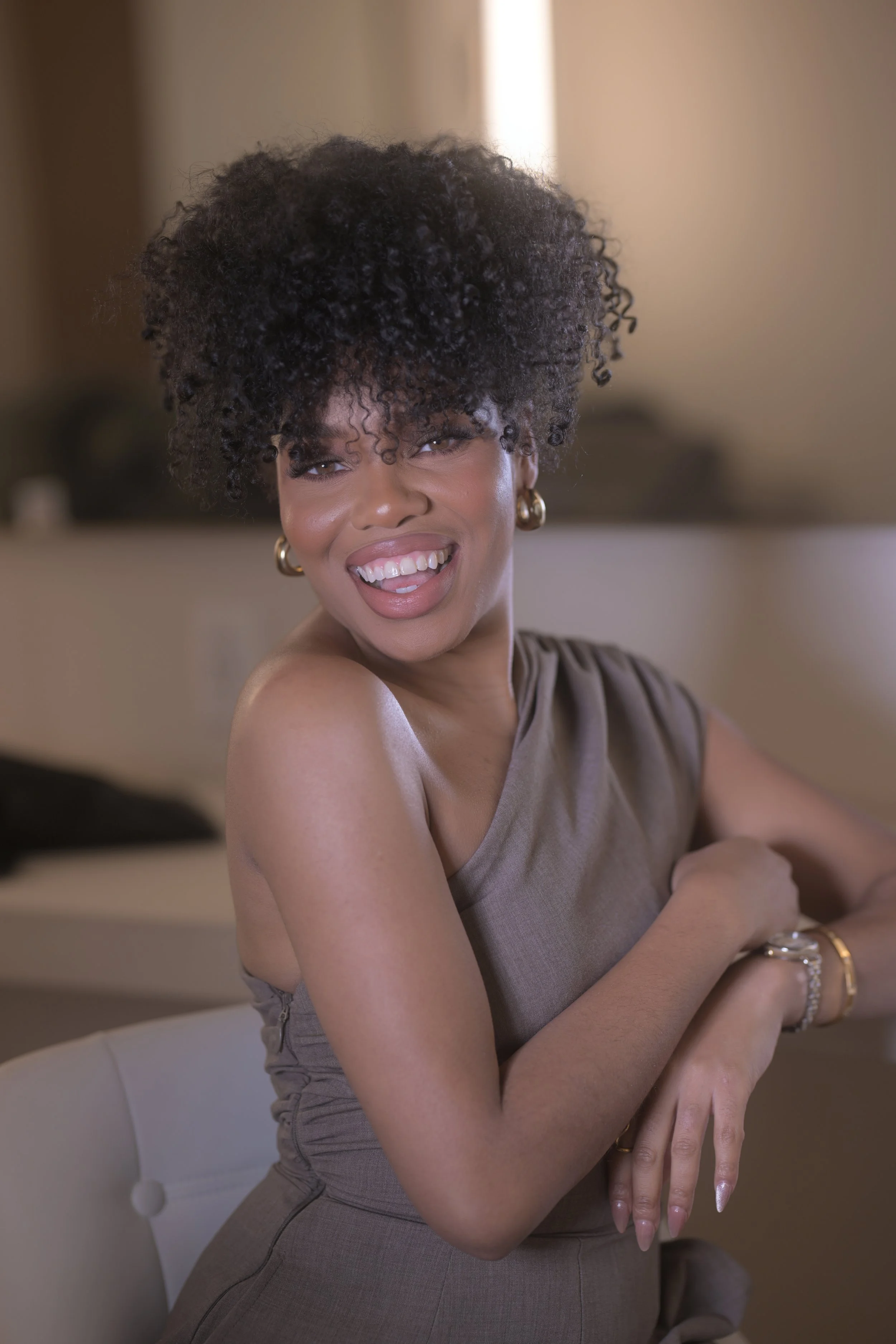 A woman smiling with her tongue slightly out, sitting on a chair indoors, wearing a sleeveless brown dress and gold jewelry.