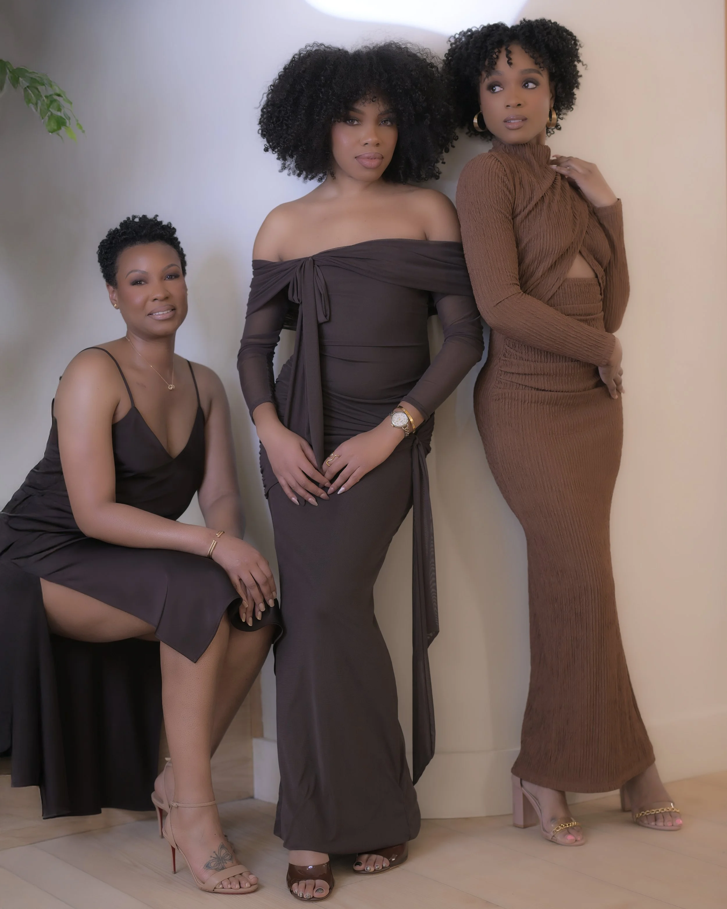 Three women dressed elegantly in dark and brown outfits, posing indoors against a light-colored wall with a green plant partially visible in the background.