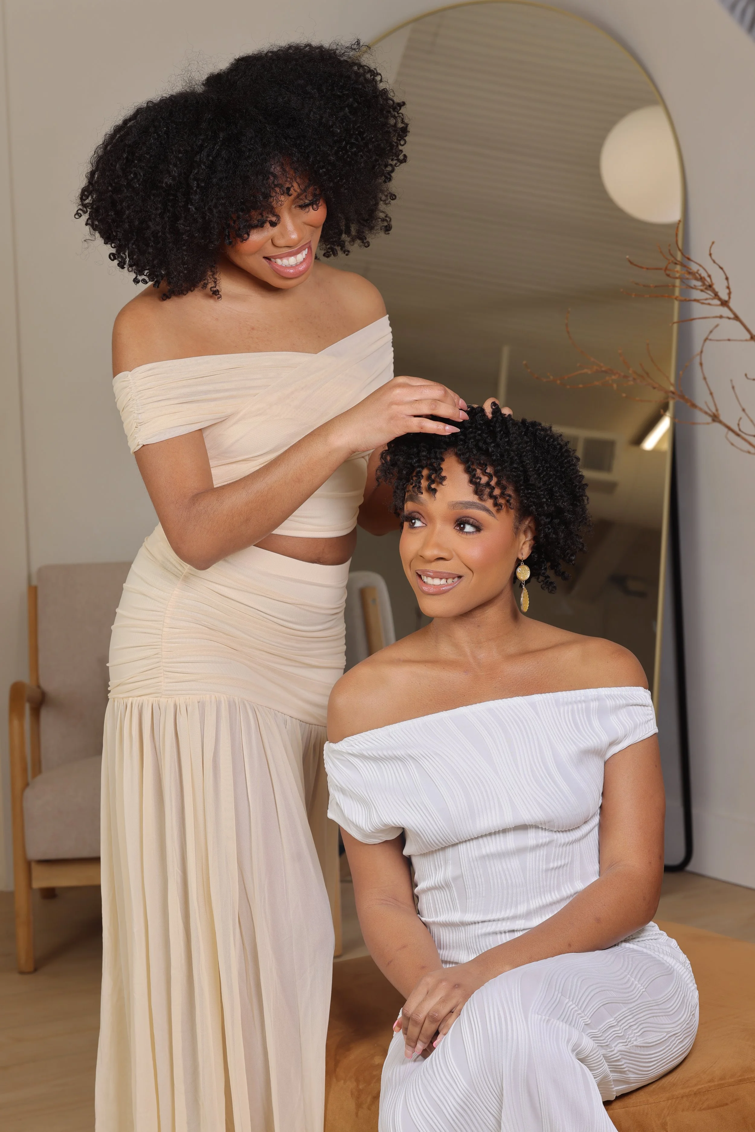 Two women with curly hair, wearing elegant off-the-shoulder dresses, one seated and one standing, in a bright room with a large mirror and a decorative branch.