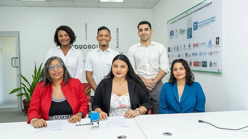 Seis personas en una reunión de oficina, con un fondo de cartel con logotipos y palabras en español, dos sentadas al frente y cuatro de pie detrás. Todos son adultos, hombres y mujeres, algunos con gafas, vestidos formalmente.