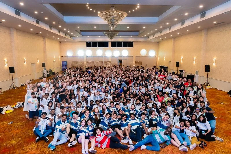 Gran grupo de personas en un salón grande, con decoraciones elegantes y luces, posando para una foto grupal.