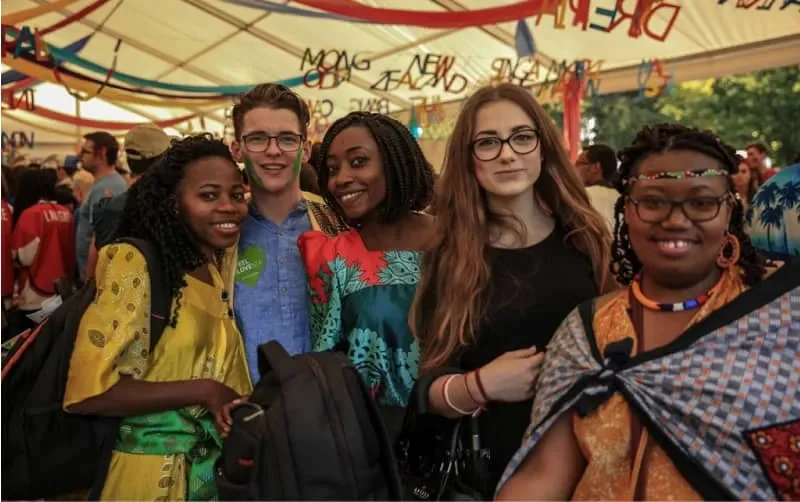 Grupo de cinco jóvenes sonriendo, en un evento al aire libre con decoraciones coloridas y muchas otras personas en el fondo.