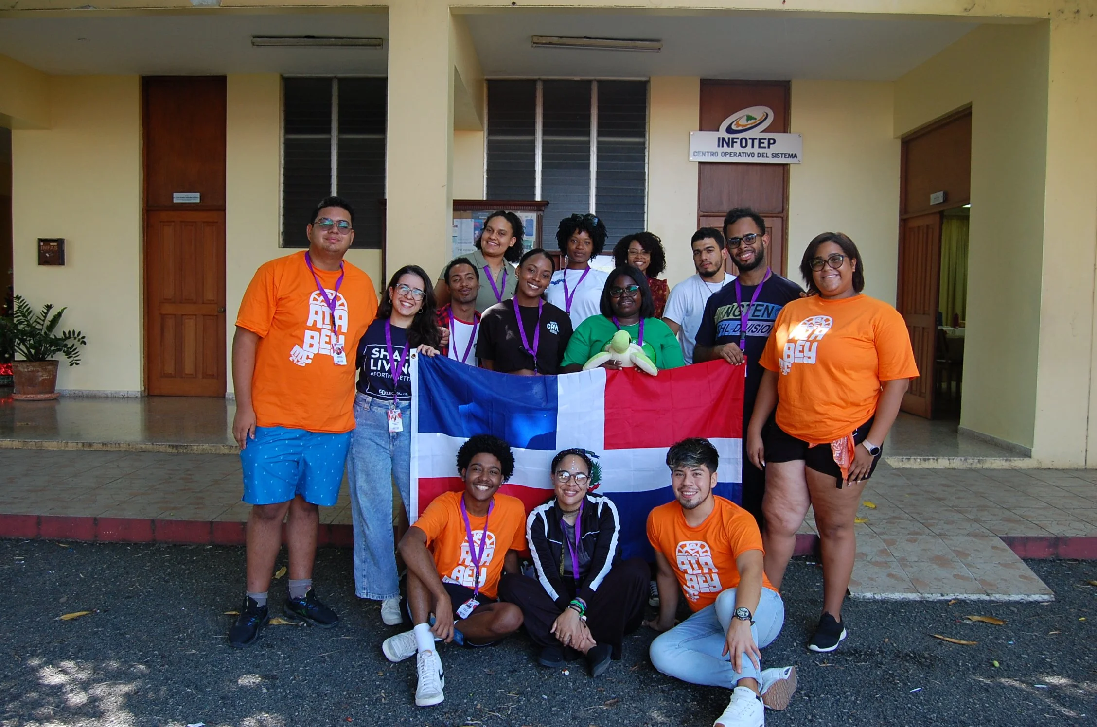 Grupo de jóvenes con medallas sosteniendo la bandera de República Dominicana frente a un edificio con un cartel que dice 'INFOTEP'.