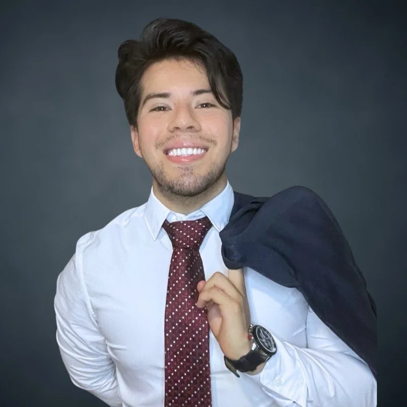Joven profesional sonriendo, con camisa blanca, corbata roja con puntos blancos, llevando saco oscuro sobre el hombro y reloj en la muñeca izquierda, fondo gris.