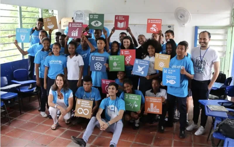 Grupo de jóvenes y adultos en un aula sosteniendo carteles relacionados con los Objetivos de Desarrollo Sostenible de la ONU.